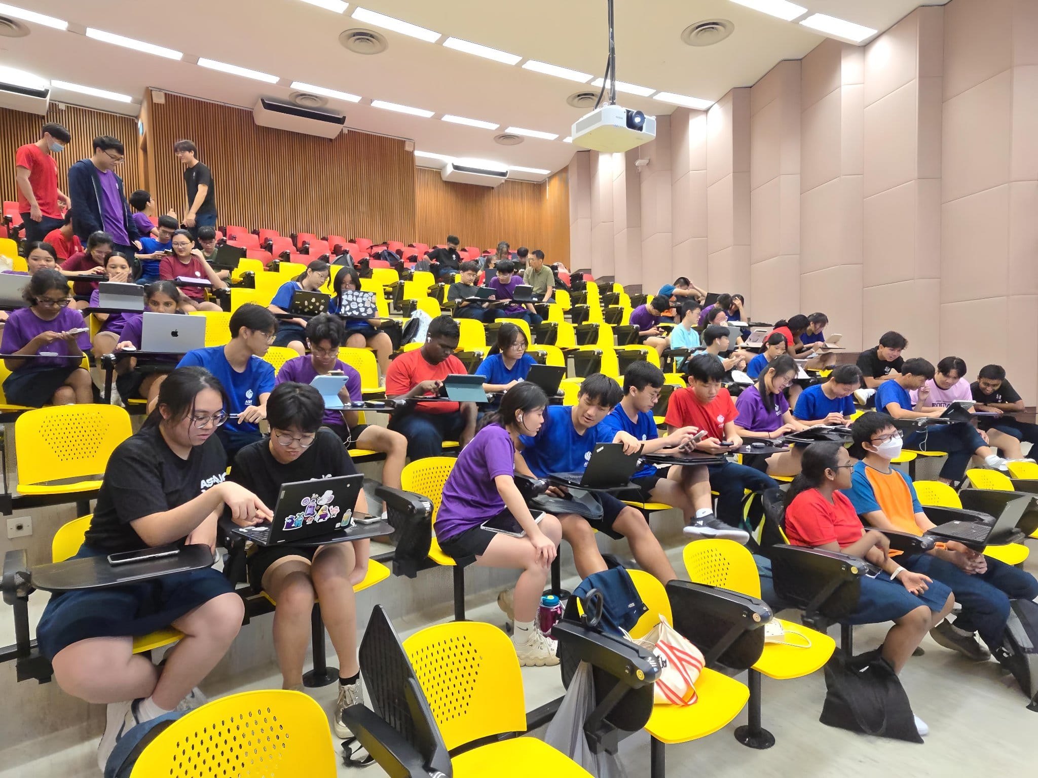 Students in colorful shirts sit in yellow lecture hall seats using laptops and tablets during a session.
