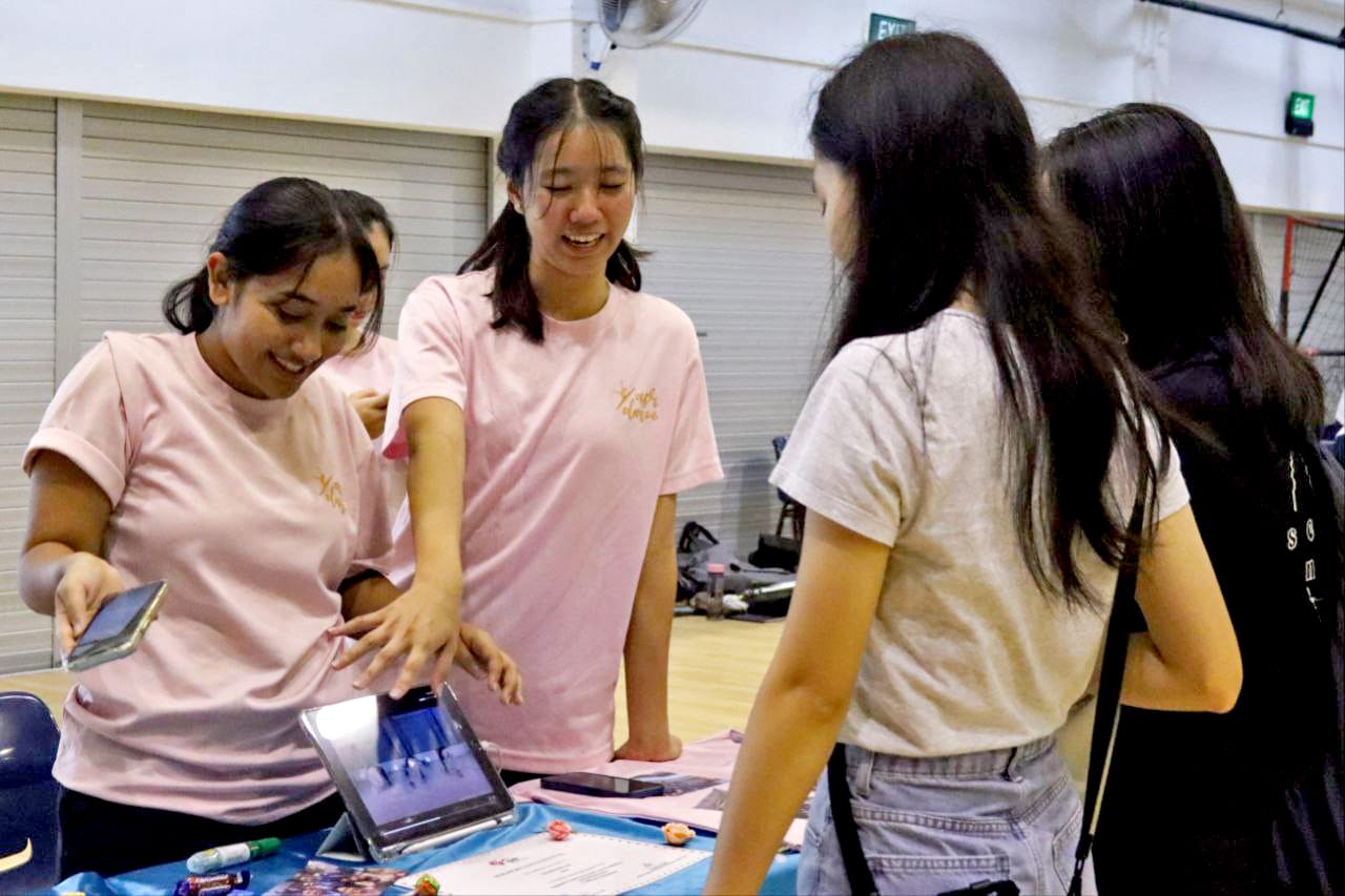 Four young women gather around a table with a tablet, two wearing matching pink shirts, smiling indoors.