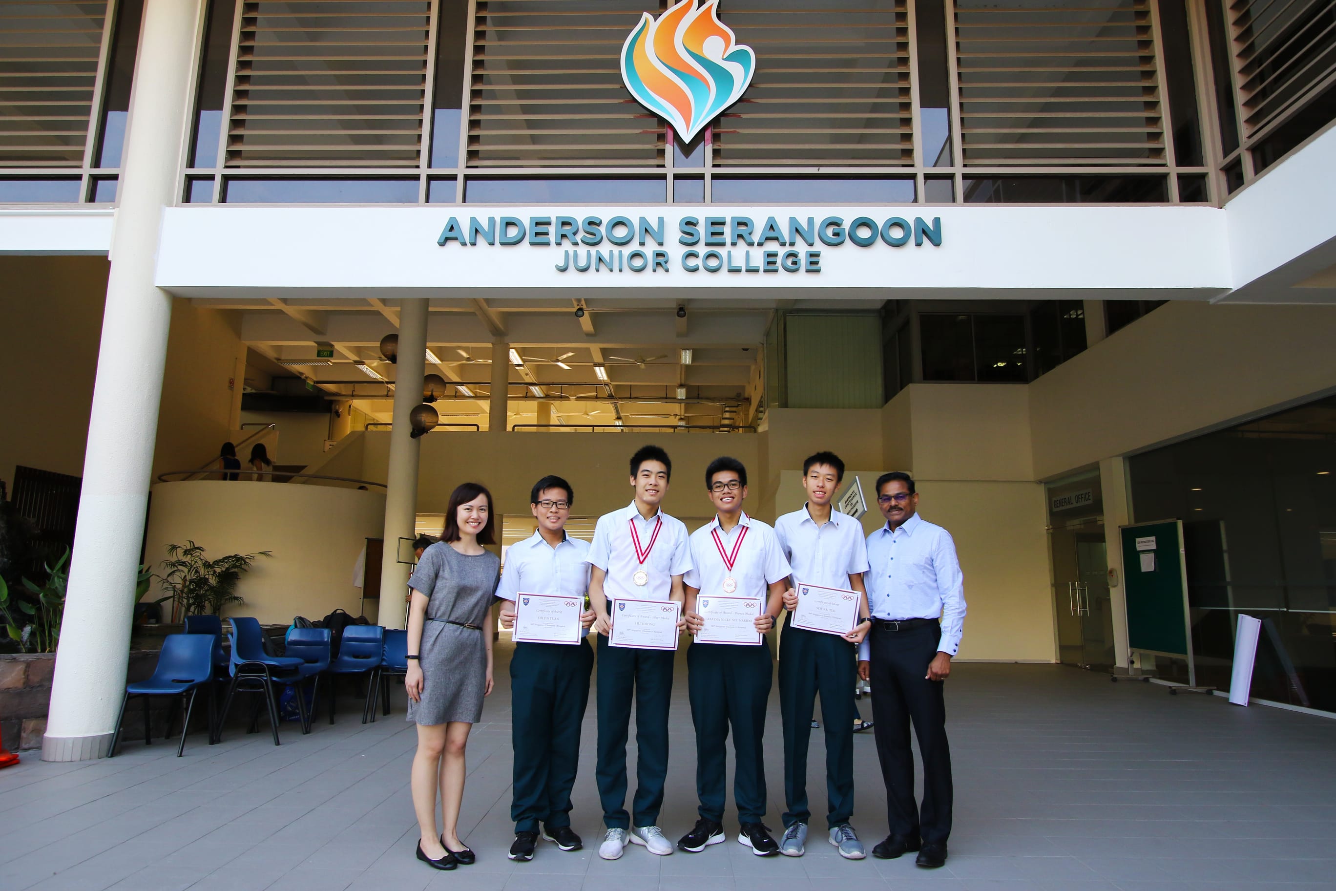 Six people stand before Anderson Serangoon Junior College; four students hold certificates and two wear medals.