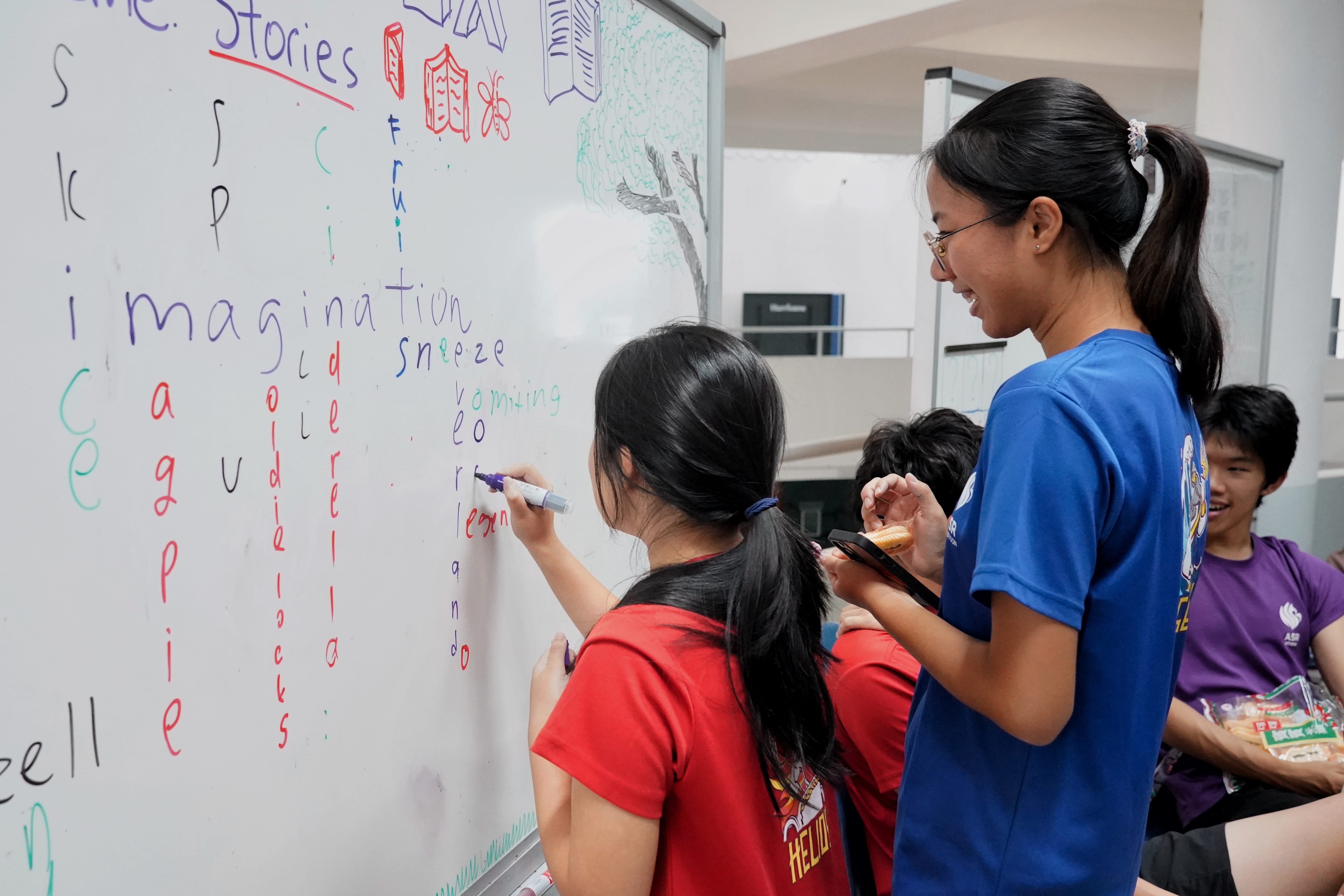 Two students write colorful words like "imagination" and "sneeze" on a whiteboard while others watch nearby.