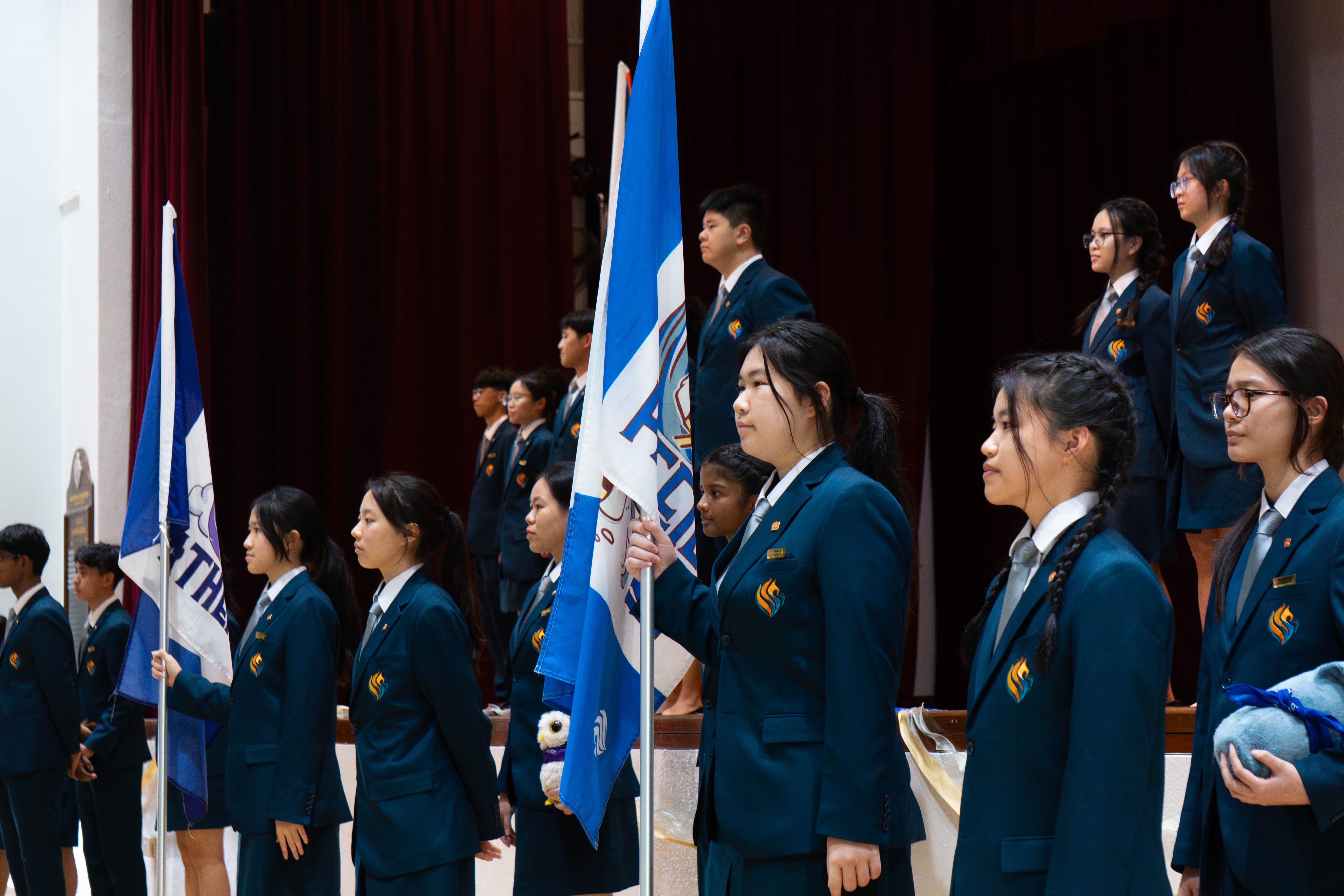 Students in dark blue uniforms stand in rows holding blue and white flags during a school ceremony.