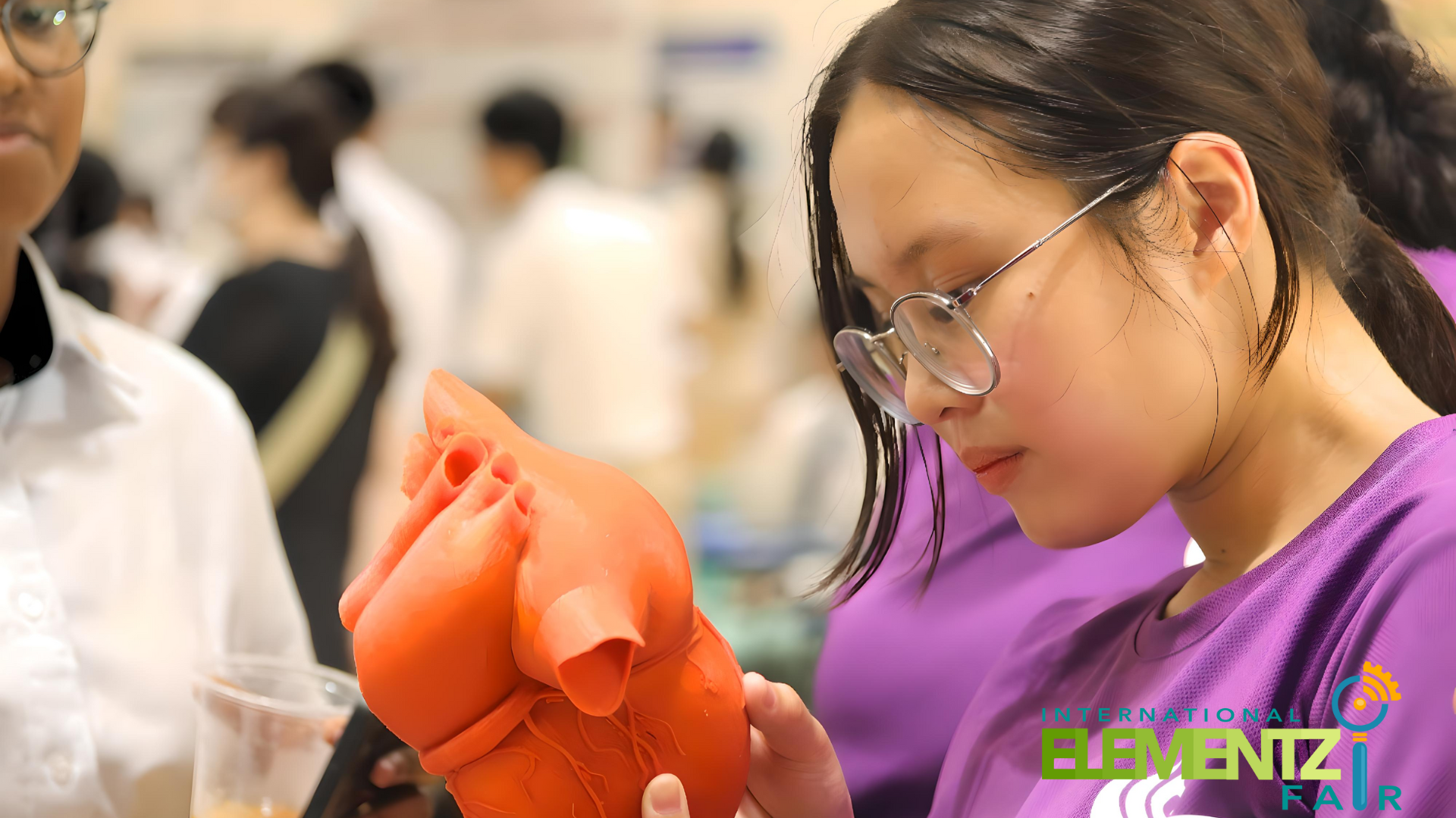 A young girl with glasses in a purple shirt closely examines a red 3D-printed heart model at a science fair.