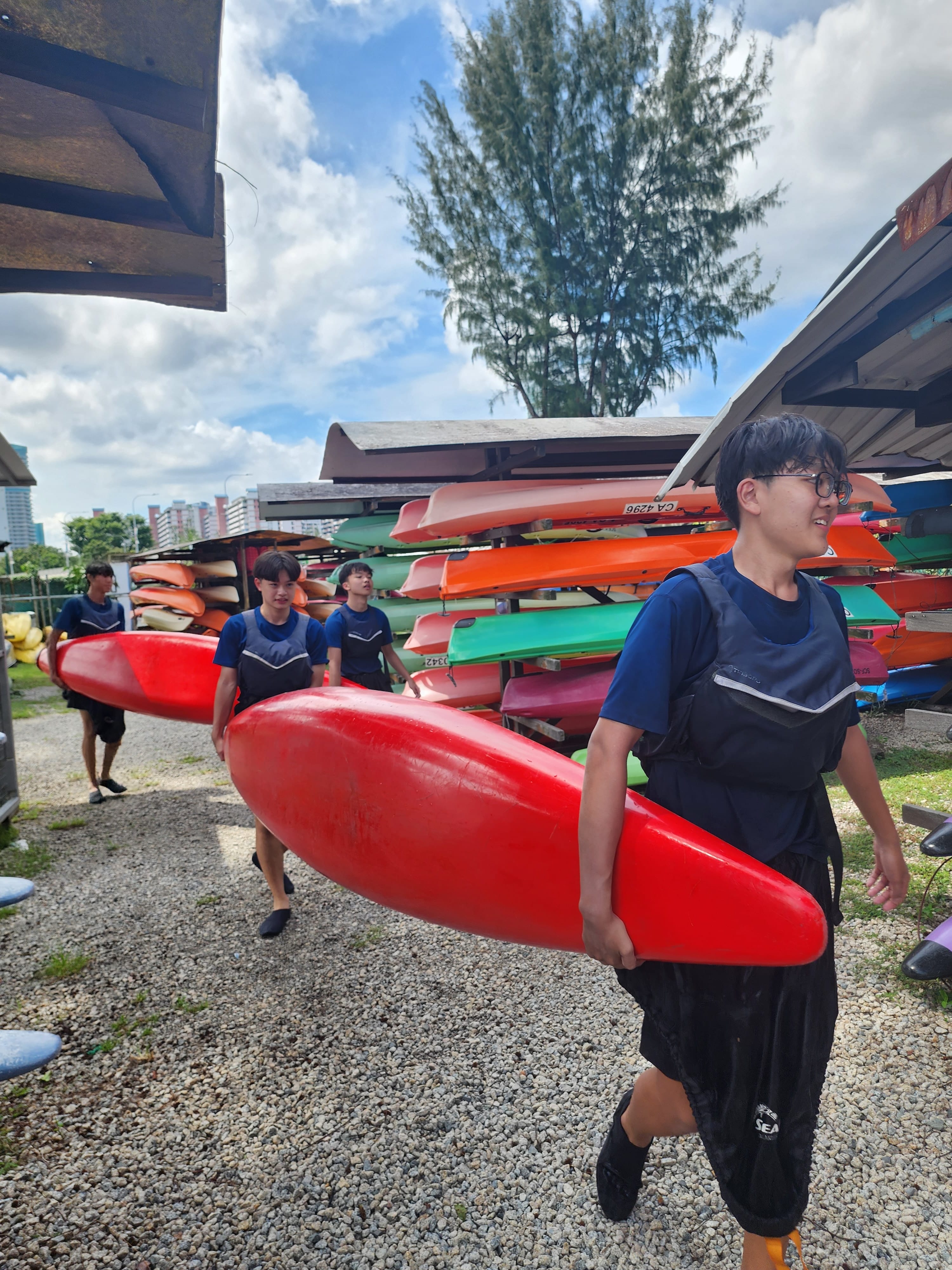 Four young people in blue shirts carry red kayaks past racks of colorful kayaks on a gravel outdoor storage area.