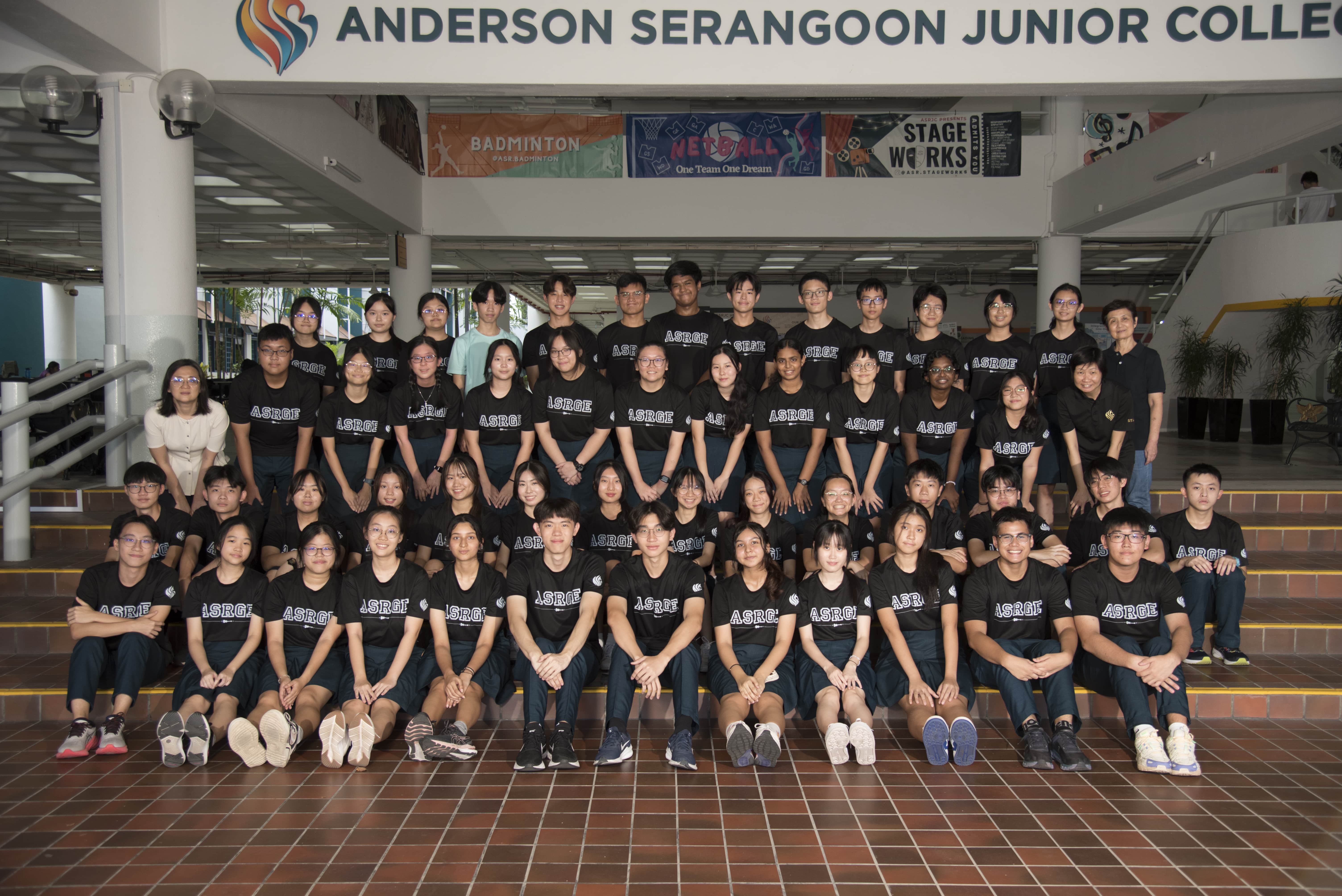 A large group of students in black ASRGE shirts and a teacher in beige top pose on steps at Anderson Serangoon Junior College.