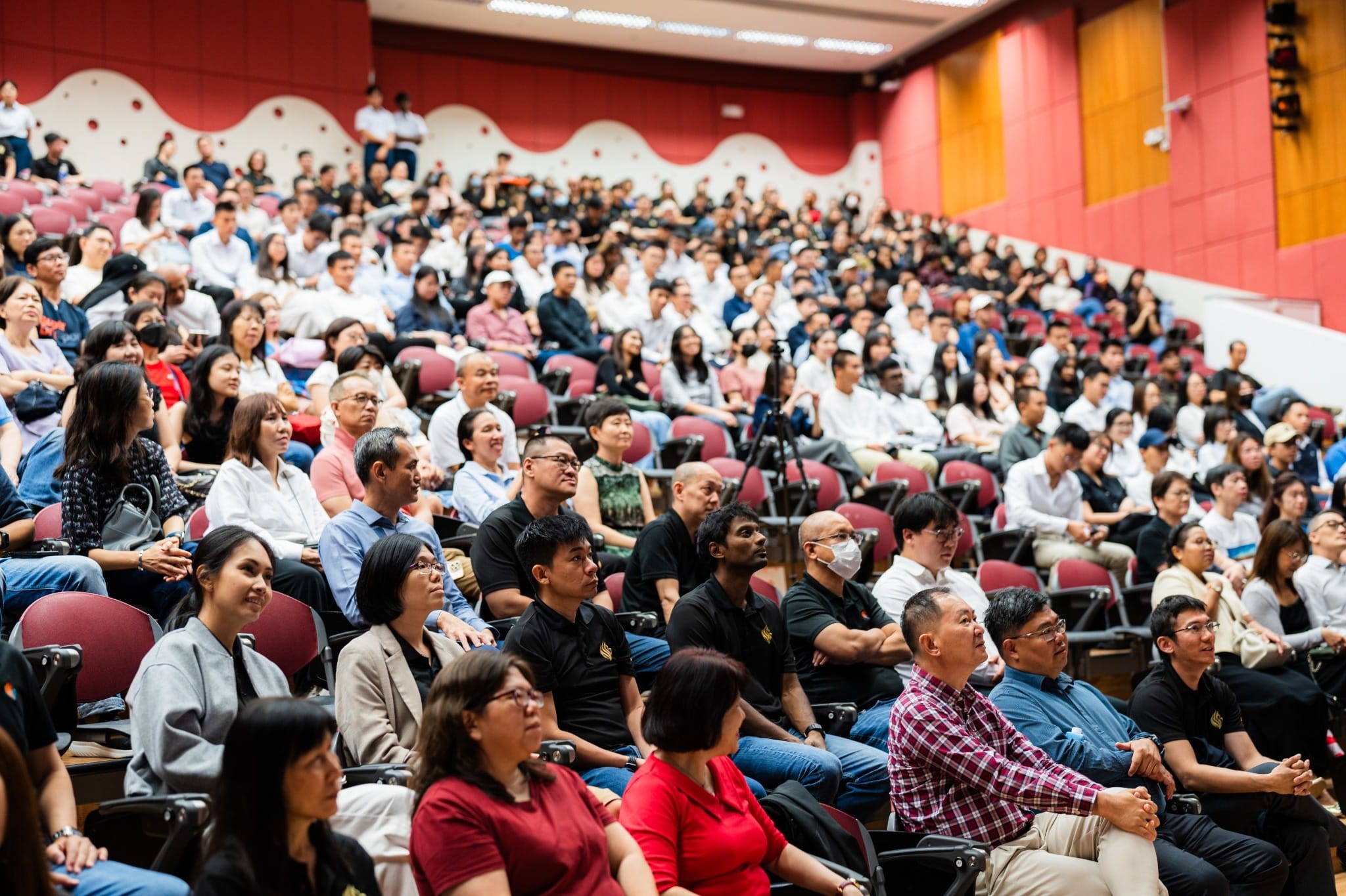 A large audience of diverse people seated in a red-walled auditorium, attentively watching an event.