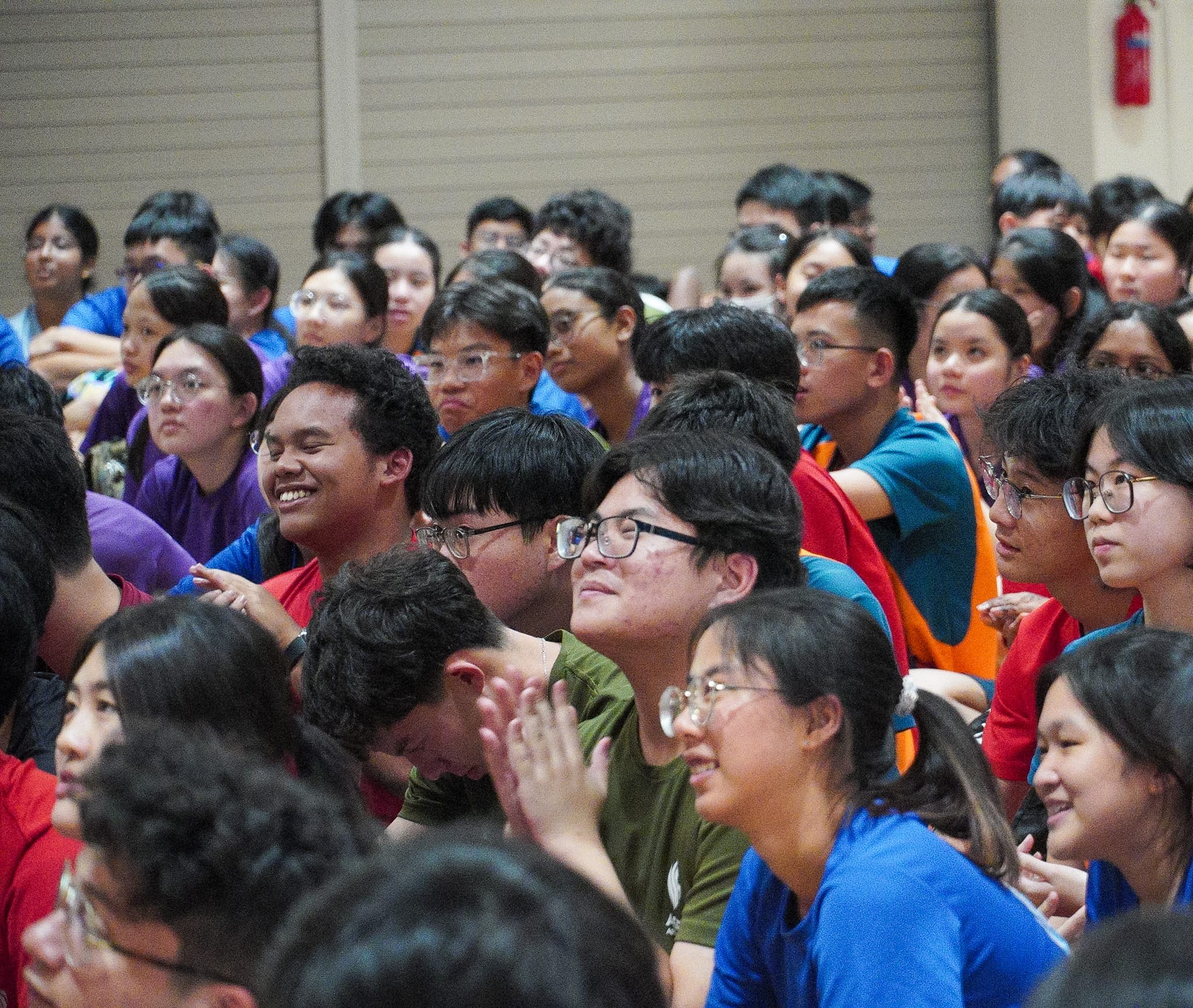 A large crowd of young Asian students in colorful shirts sitting together, many smiling and clapping at an event.
