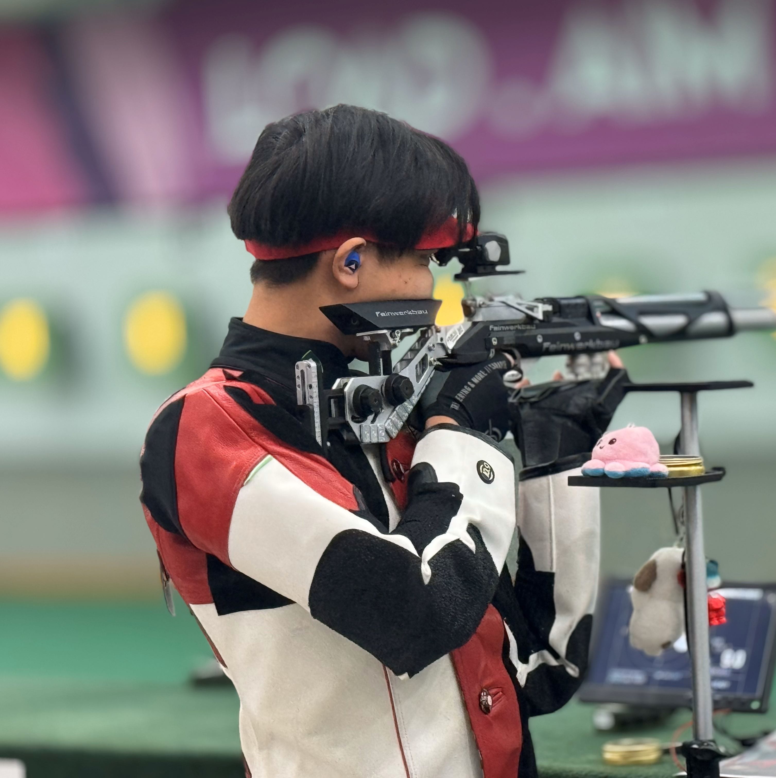 A competitive shooter in a red and white jacket aims a precision air rifle at an indoor shooting range.