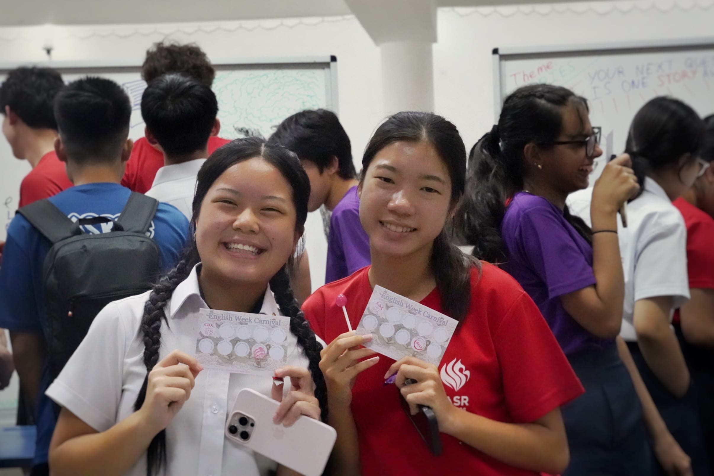 Two smiling teenage girls hold English Week Carnival cards in a busy classroom with whiteboards and students in the background.