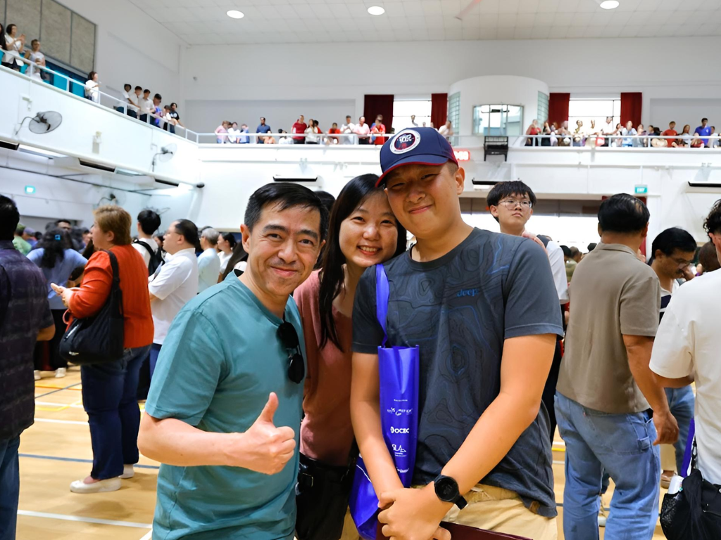 Three smiling people pose in a crowded indoor hall, one giving a thumbs up, another holding a blue bag.