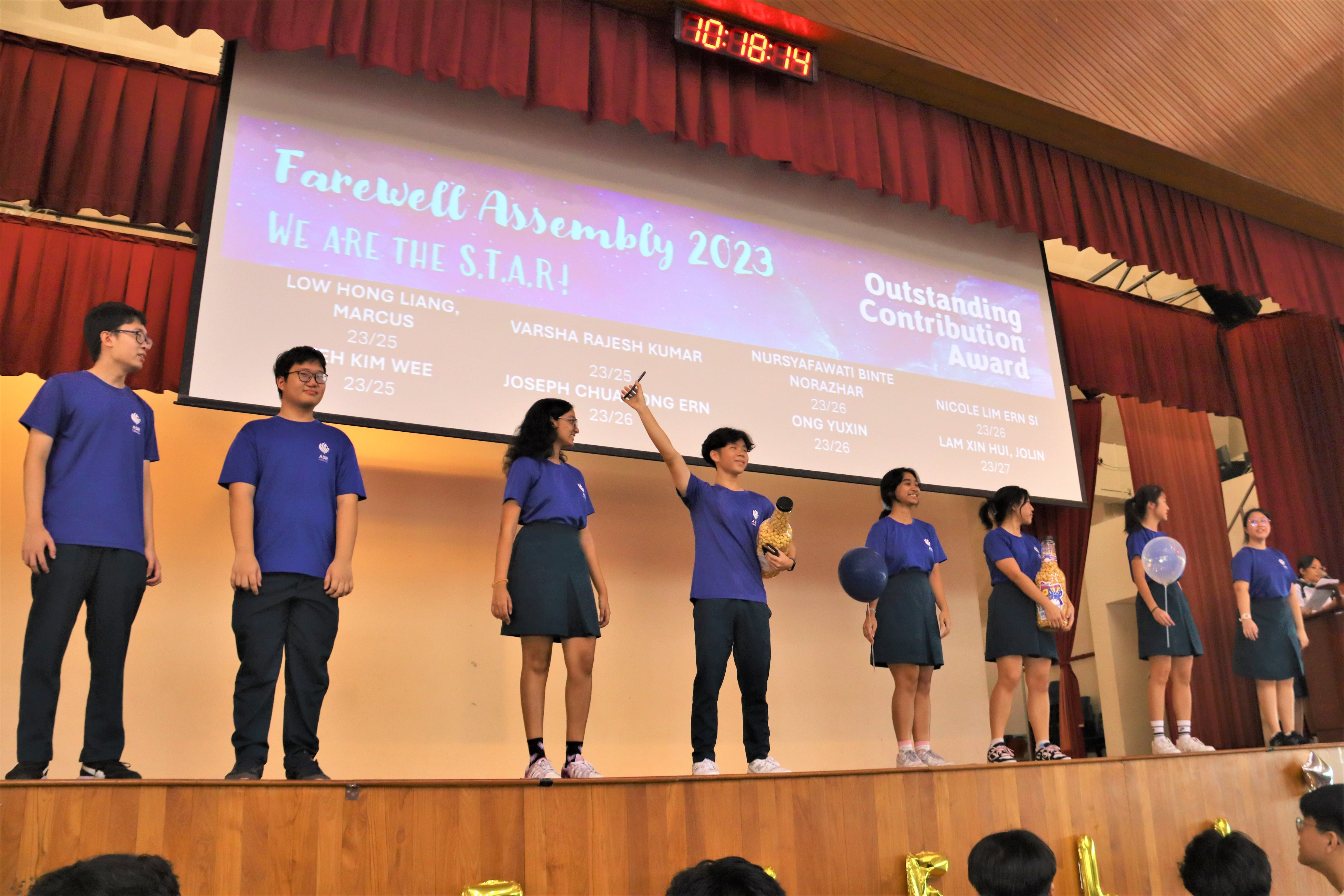 Students in blue uniforms stand on a school stage at Farewell Assembly 2023 with an Outstanding Contribution Award slideshow behind them.