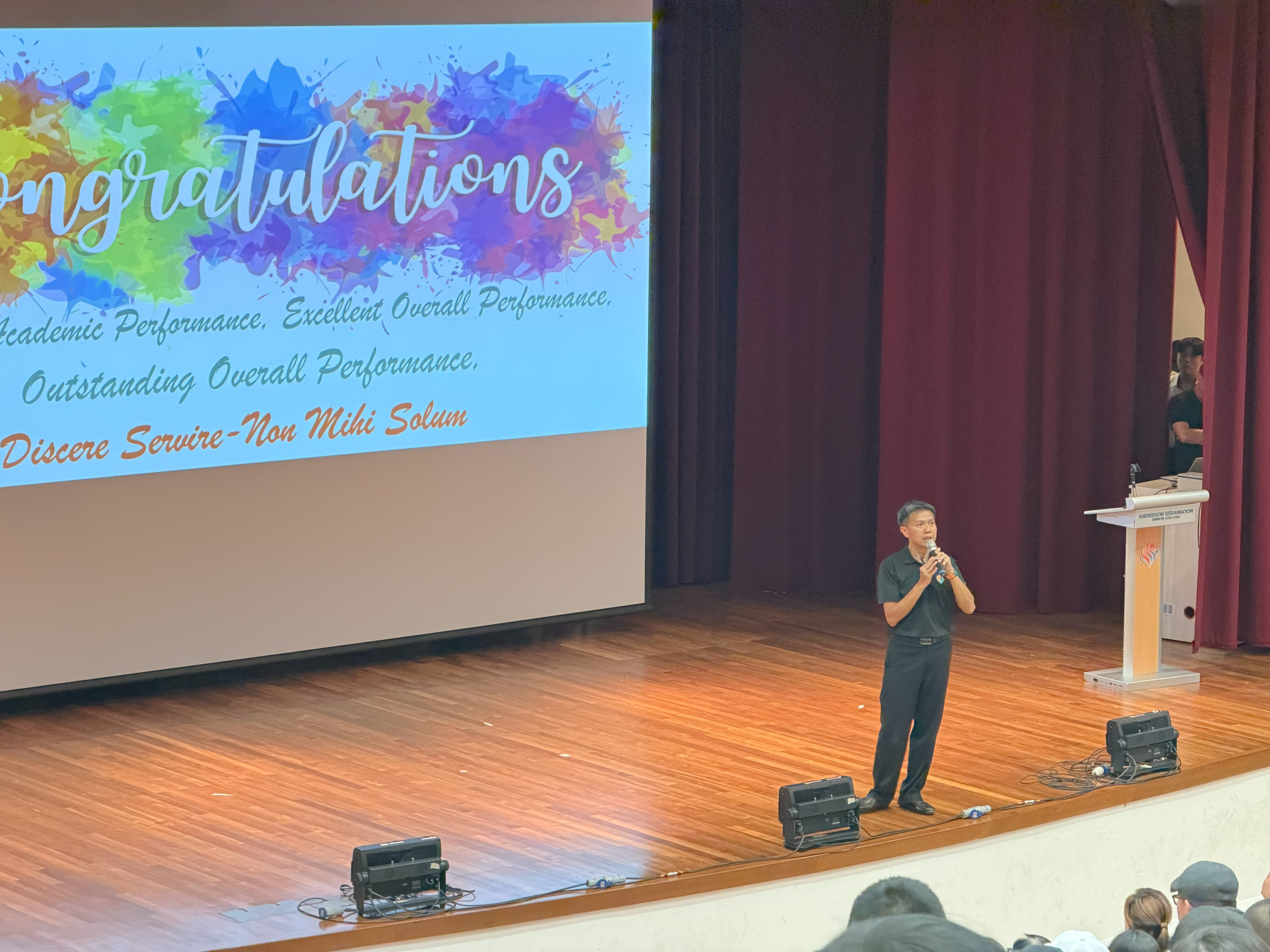 A man in black stands on the stage to speak to the audience in front a Congratulations slide
