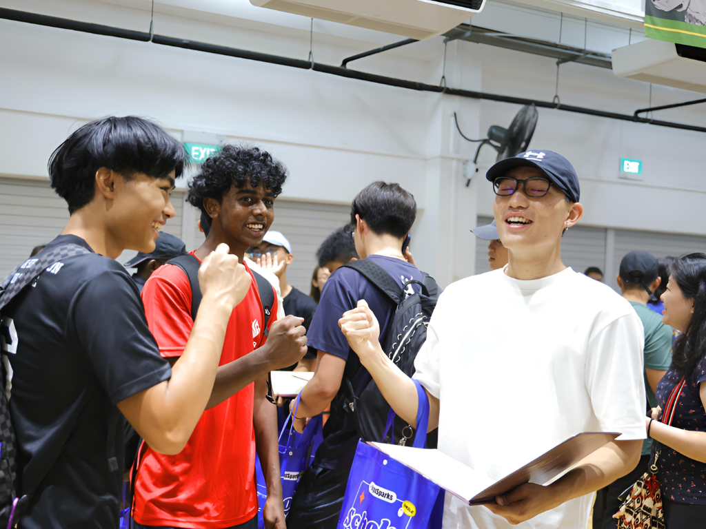A group of smiling young people interact indoors, one wearing a white shirt and blue cap holding a folder and bag.
