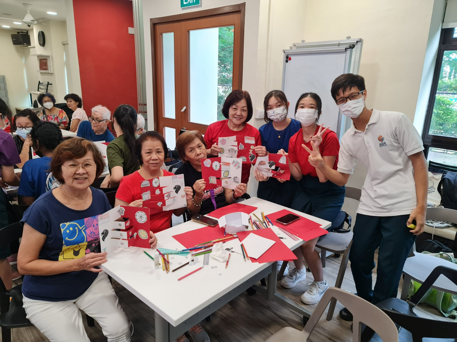 A group of seniors and young volunteers smile and hold up handmade red cards at a community craft activity indoors.