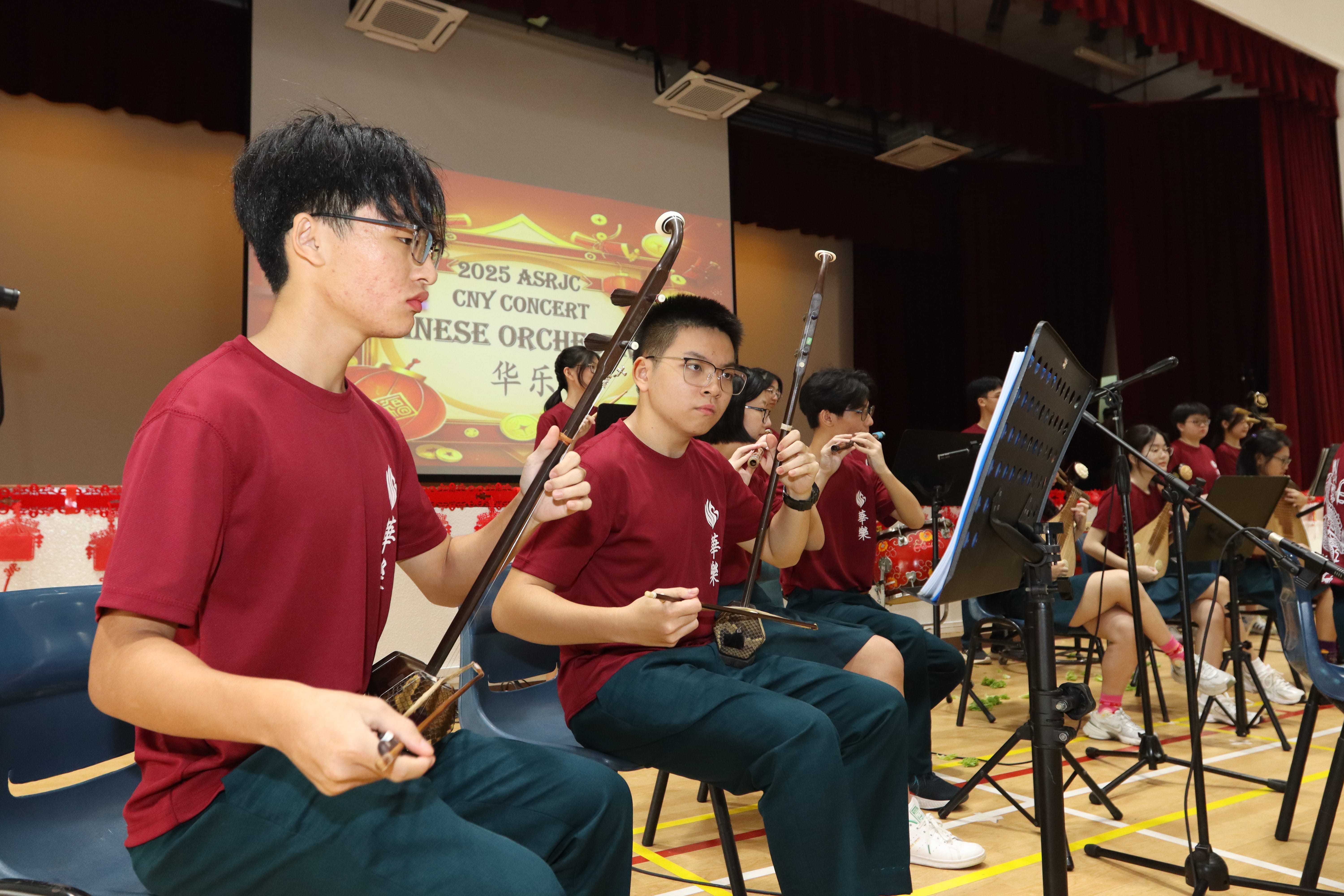 Students in maroon shirts play erhu and other instruments on stage, reading sheet music on stands.