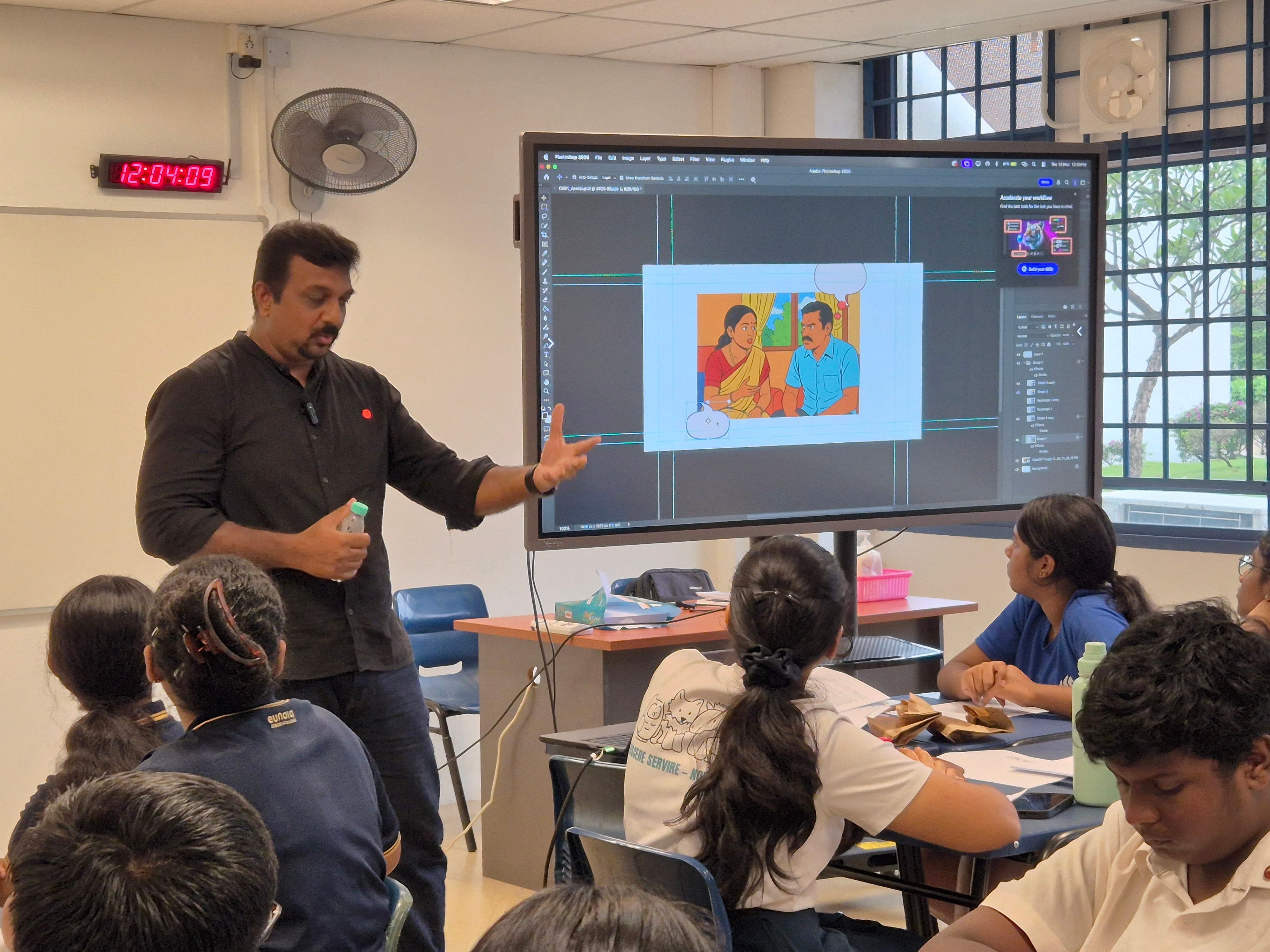 A man in black presents a Photoshop illustration on a large screen to students seated in a classroom.