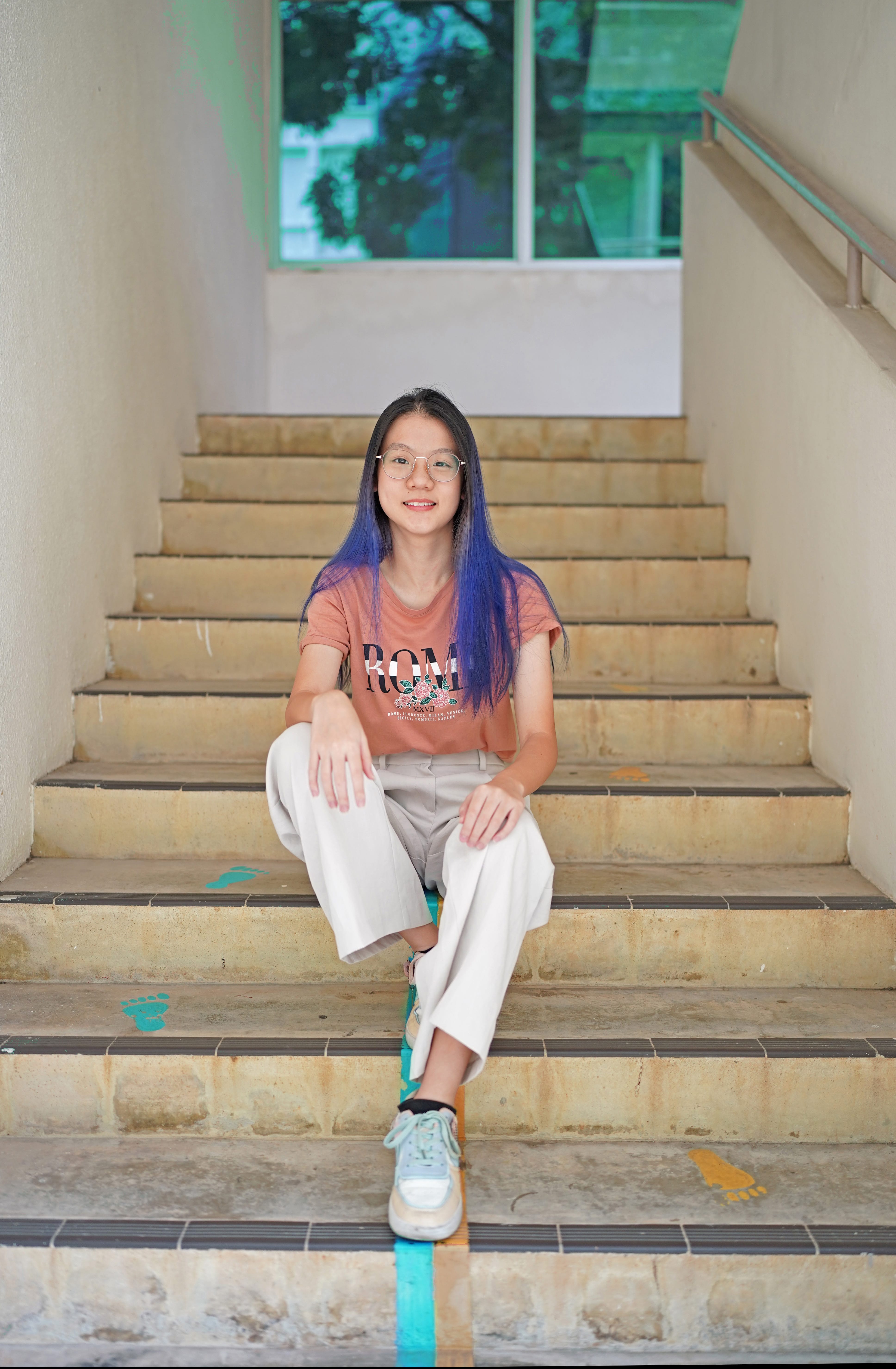 Young woman with blue-dyed hair and glasses sits smiling on concrete stairs wearing a pink tee and white pants.
