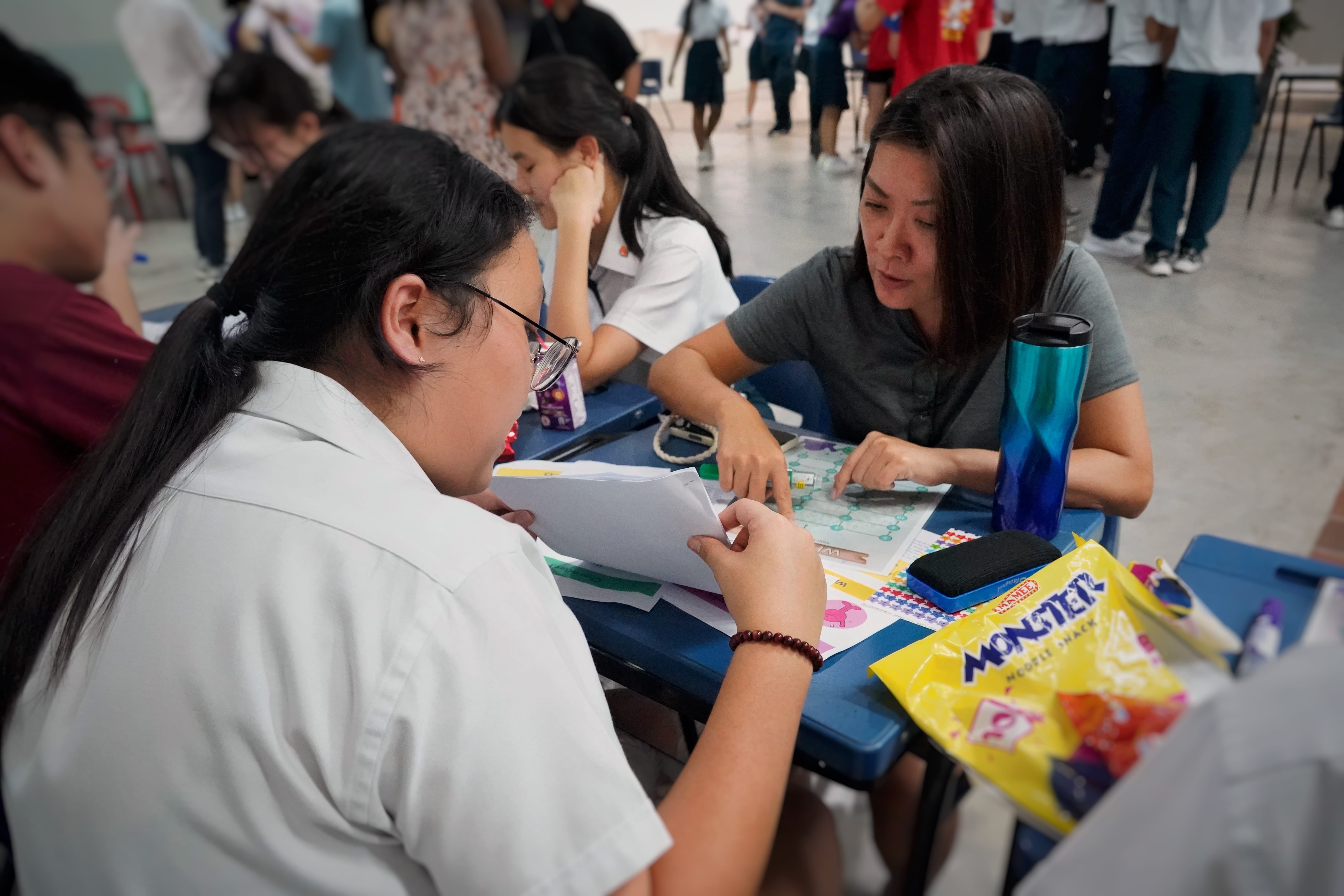 Teacher points at a board game on a classroom desk as a student reads papers; snack bag and bottle nearby.