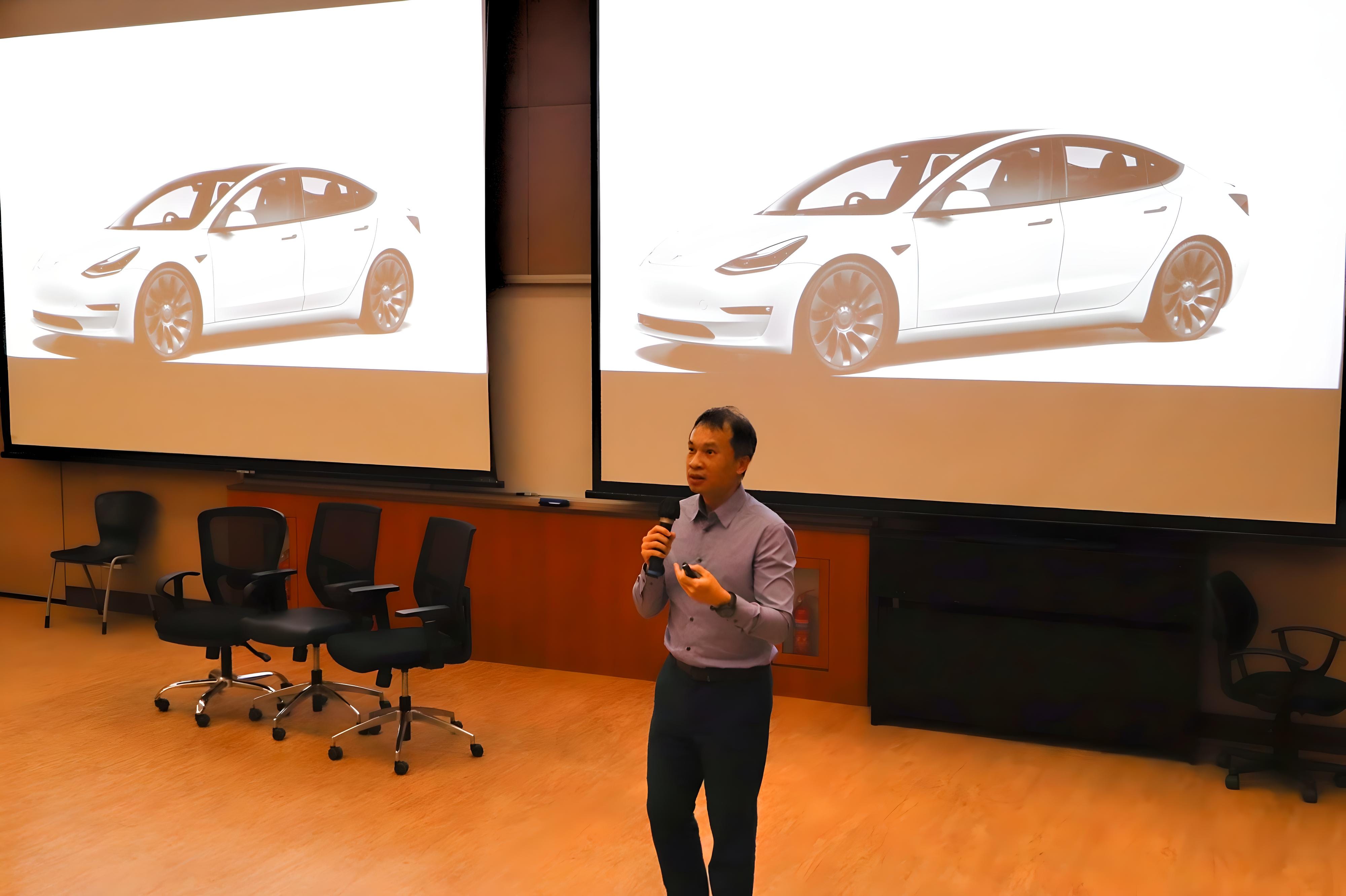 A man holds a microphone while presenting in front of two large screens showing a white Tesla car.