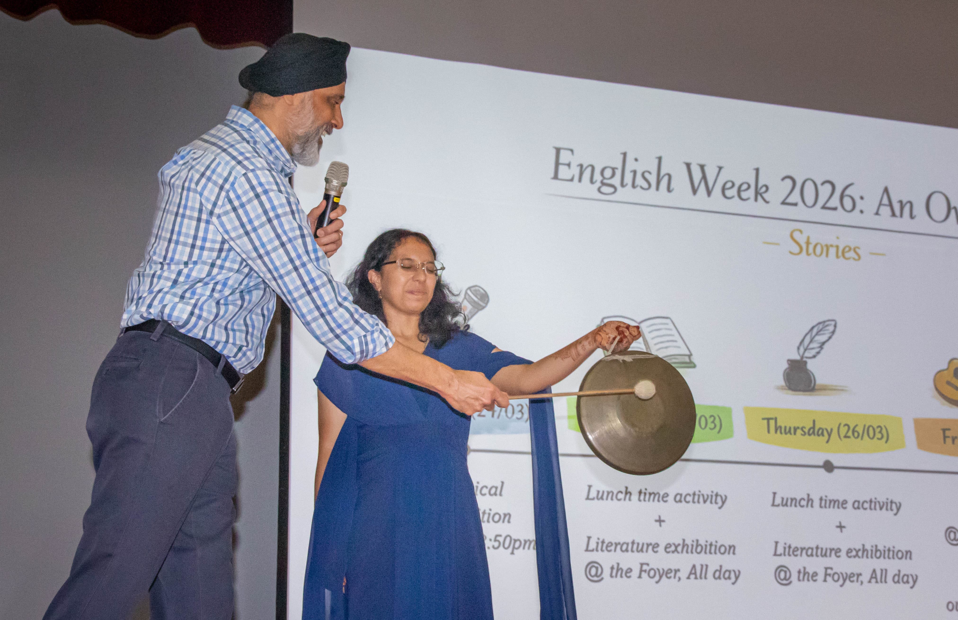 A man with a microphone and a woman in blue striking a gong in front of an English Week 2026 presentation slide.