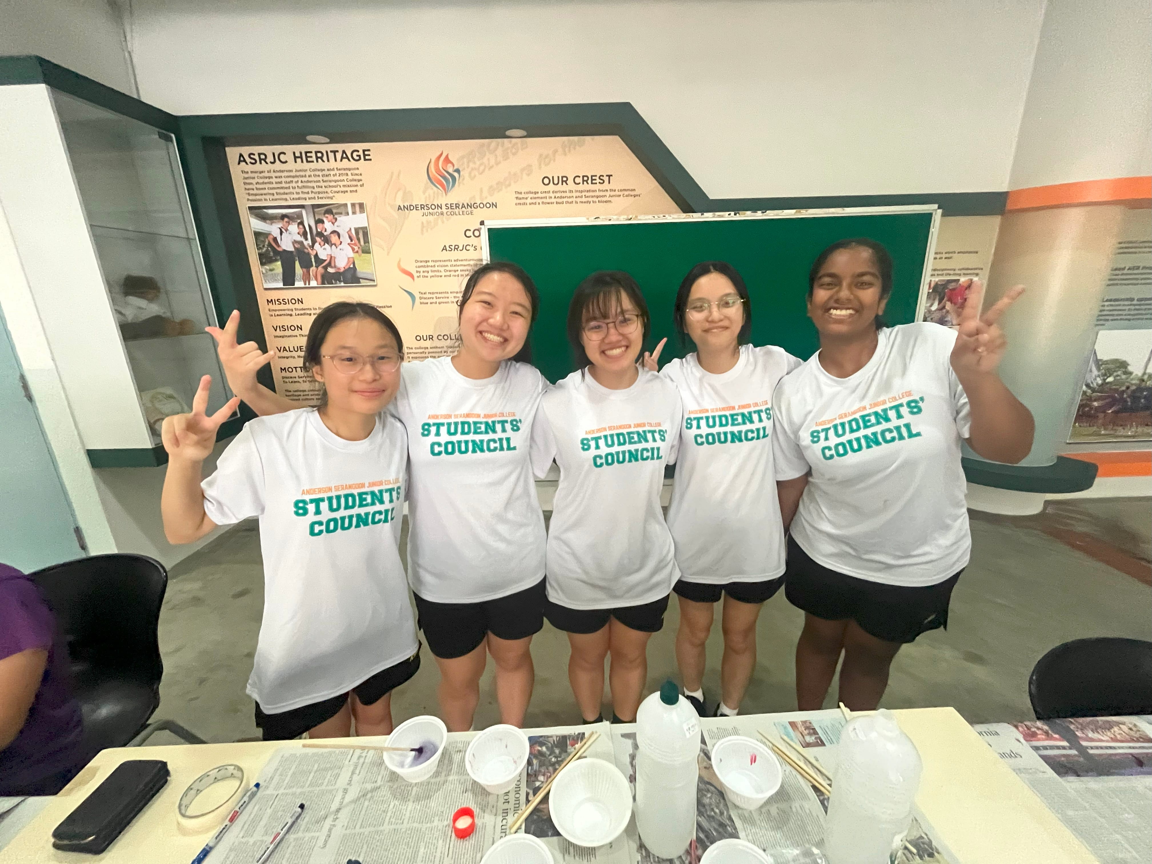 Five students wearing Anderson Serangoon Junior College Students' Council white shirts pose smiling with peace signs.