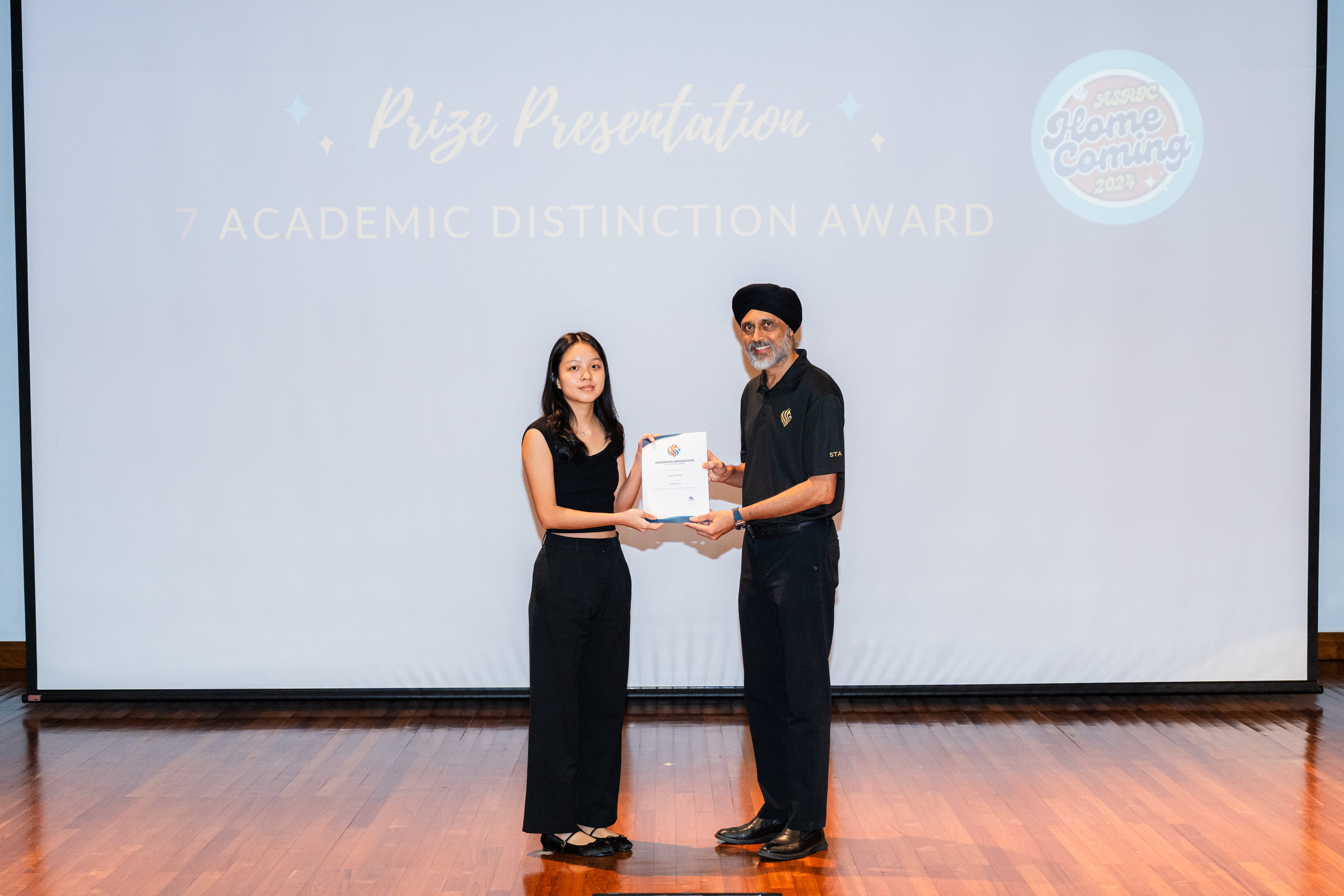 Two people in black clothing hold a certificate on stage before a screen reading Prize Presentation 7 Academic Distinction Award.