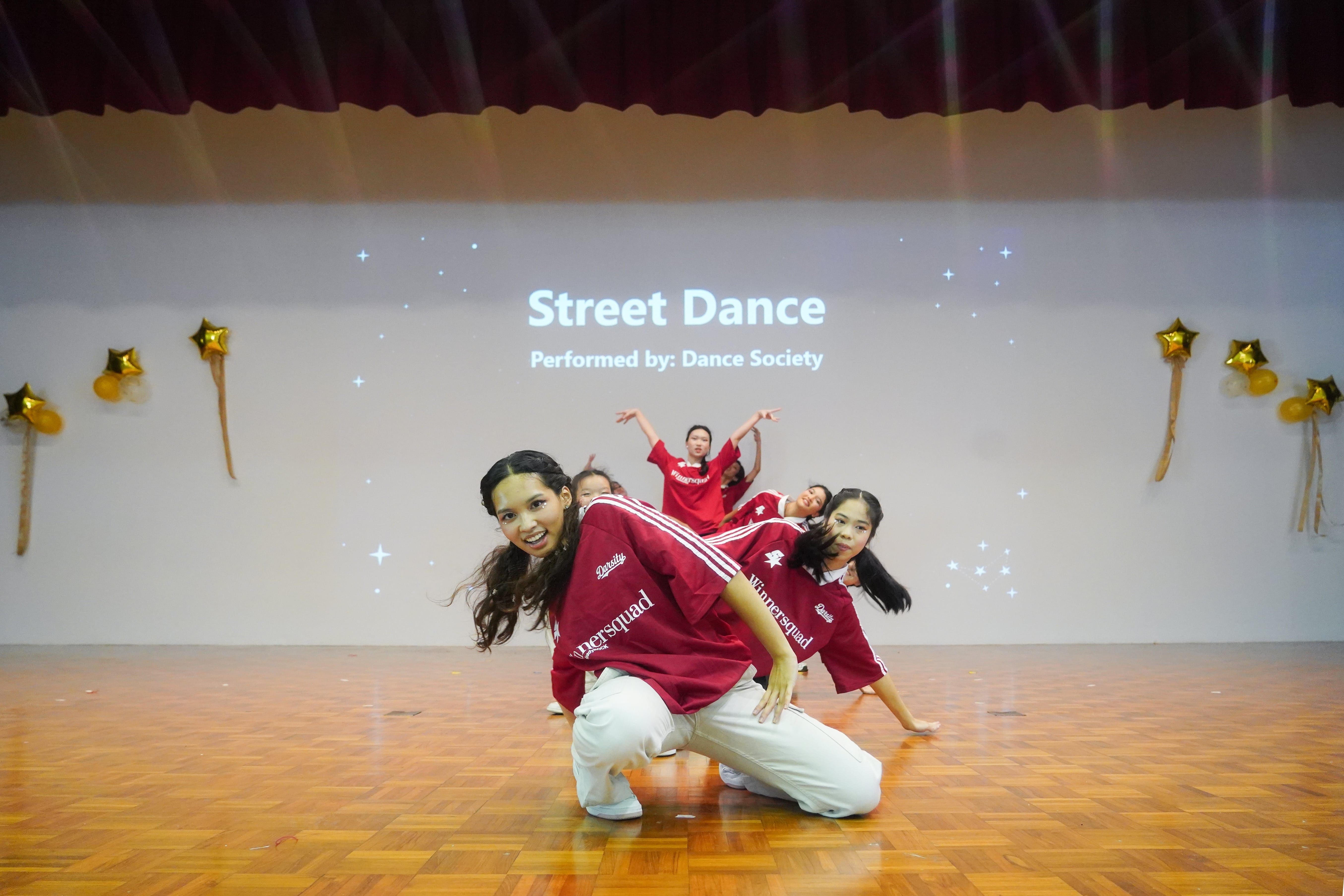 Girls in red jerseys perform street dance on a wood floor stage with a projected screen reading "Street Dance - Dance Society."