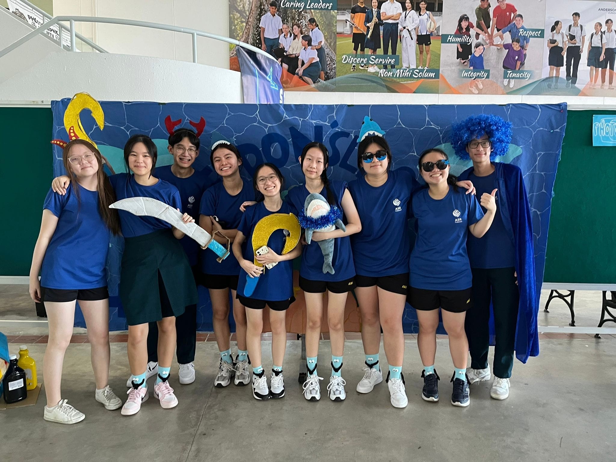 Nine students in blue shirts pose in front of a blue ocean-themed backdrop, holding props like a toy shark and sword.