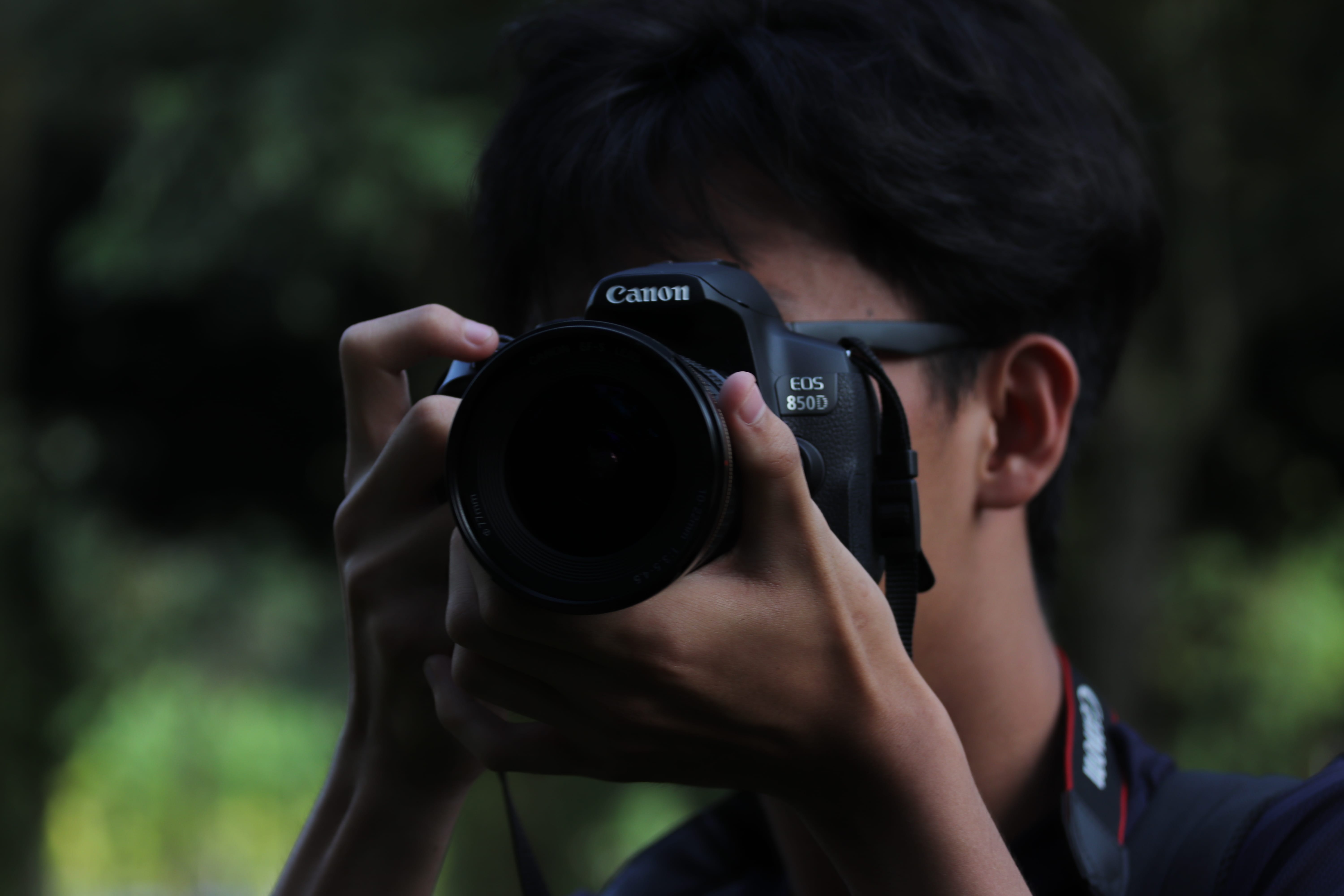 Person holding a Canon EOS 850D camera to their face, taking a photo outdoors with blurred greenery behind.
