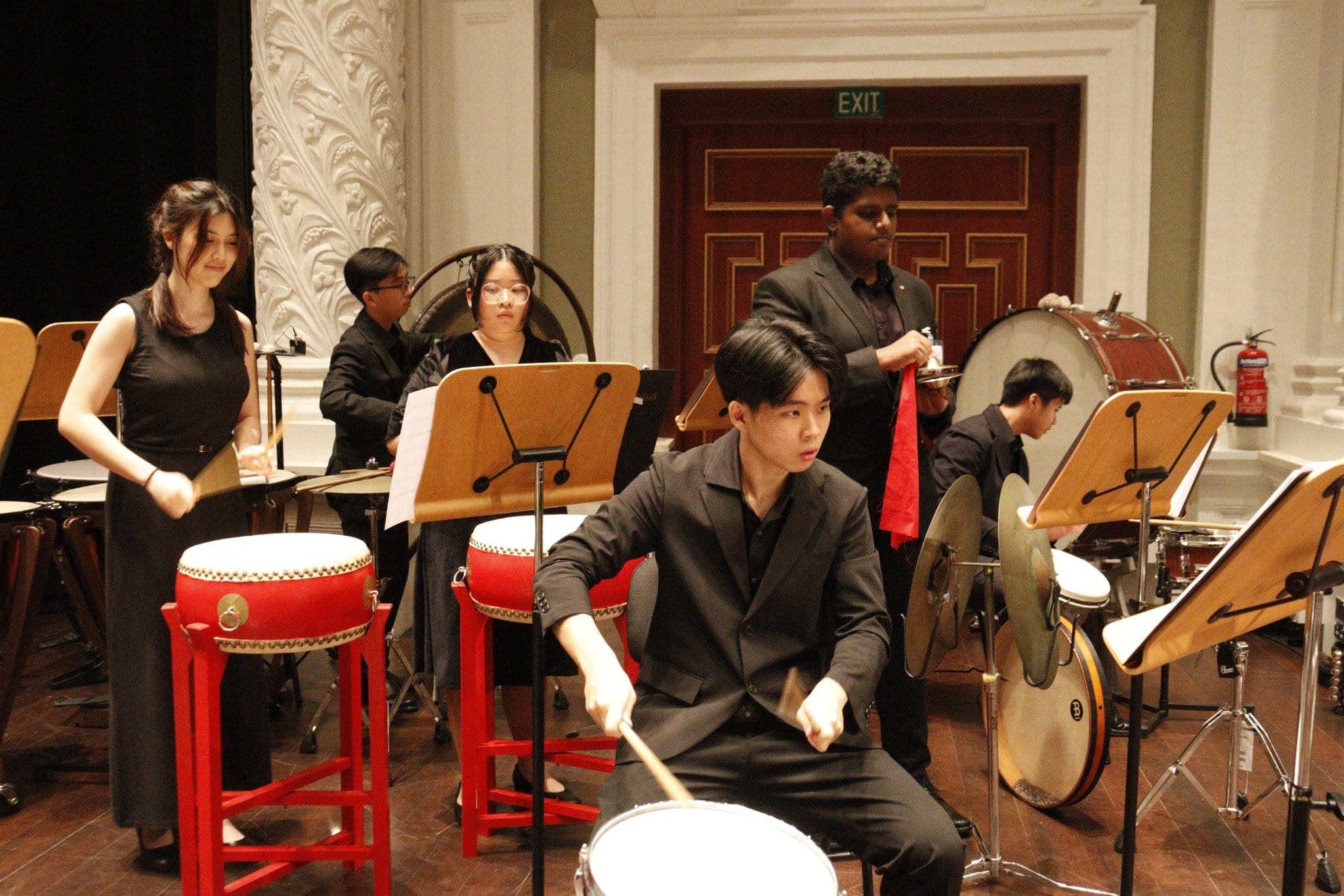 Several young musicians in black attire perform on drums, cymbals, and taiko drums in an ornate hall.