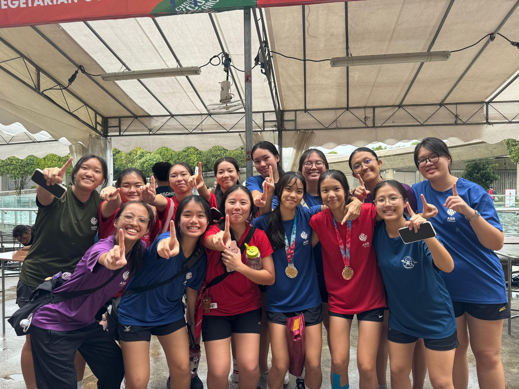 A group of young women in red and blue sports shirts pose with peace signs; two wear gold medals around their necks.