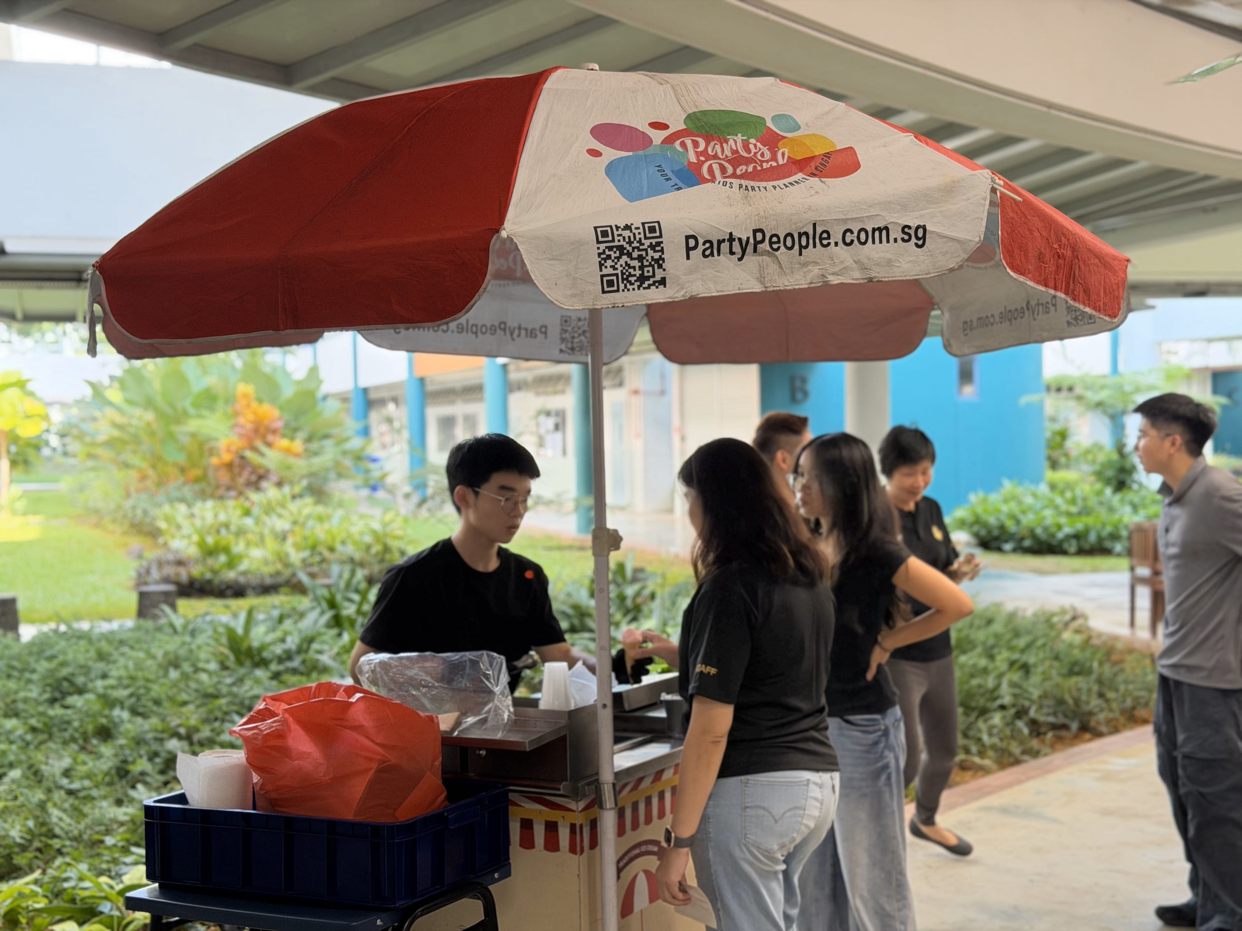2 women wait in front of an ice cream booth indoors while a man prepares the food