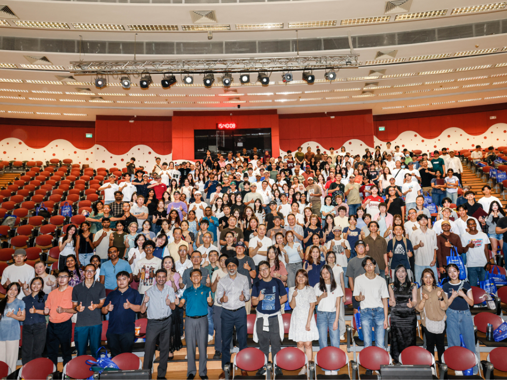 Large group of people posing with thumbs up in a red auditorium with tiered seating and stage lights.