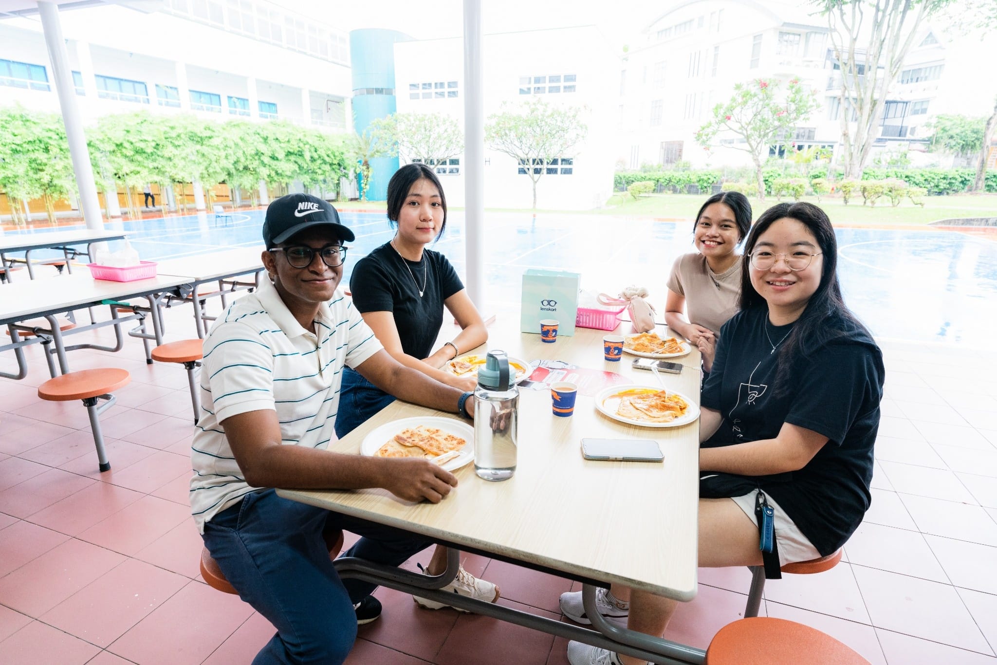 Four people smile at a table with food and drinks in an open-air school canteen with buildings and trees behind them.