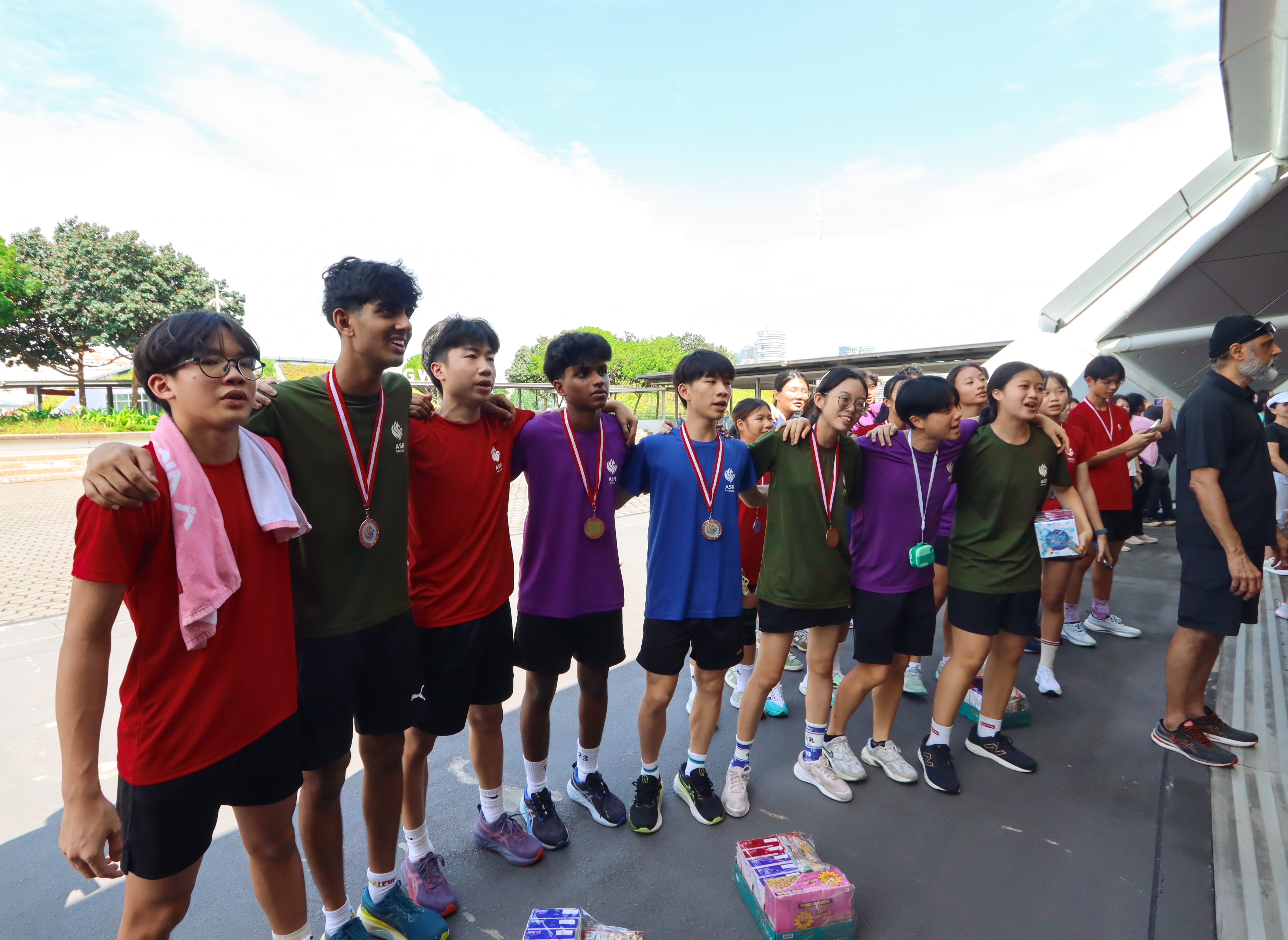 Group of students in colorful sports shirts stand arm in arm wearing medals outdoors near a building.