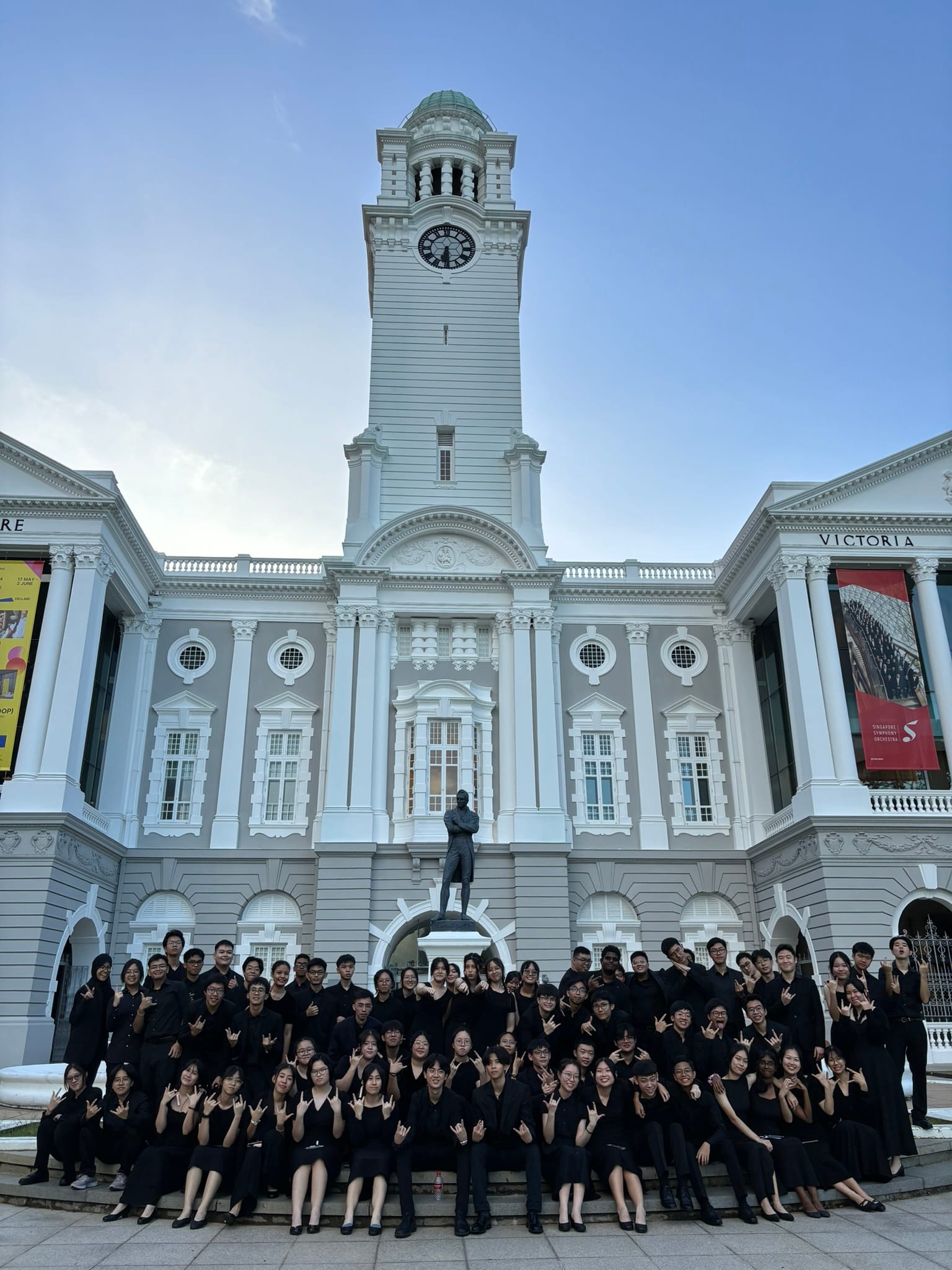 Large group of people dressed in black posing in front of Victoria Theatre with its tall clock tower in Singapore.