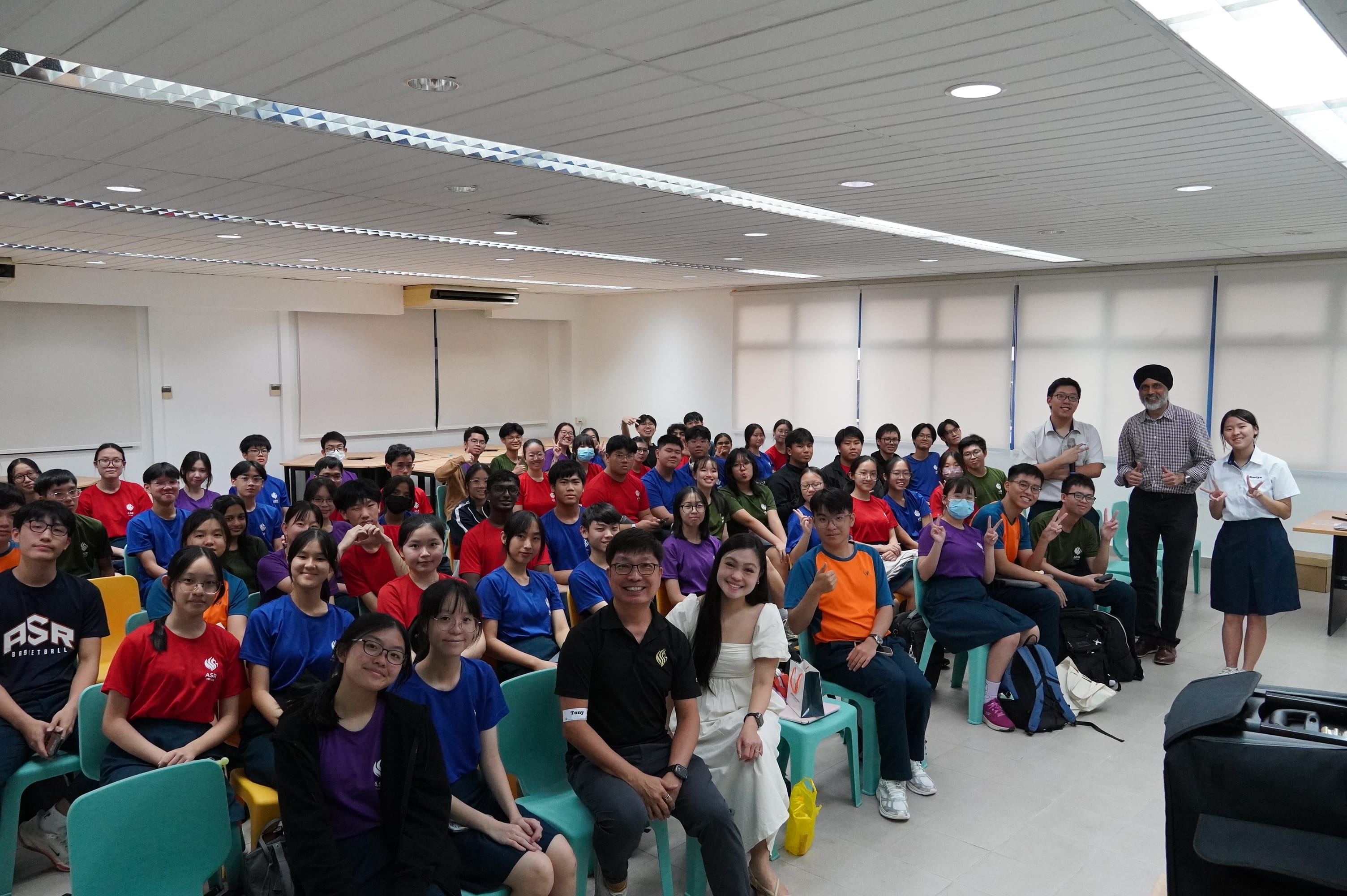 Large group of students and teachers posing together in a classroom, wearing colorful shirts.