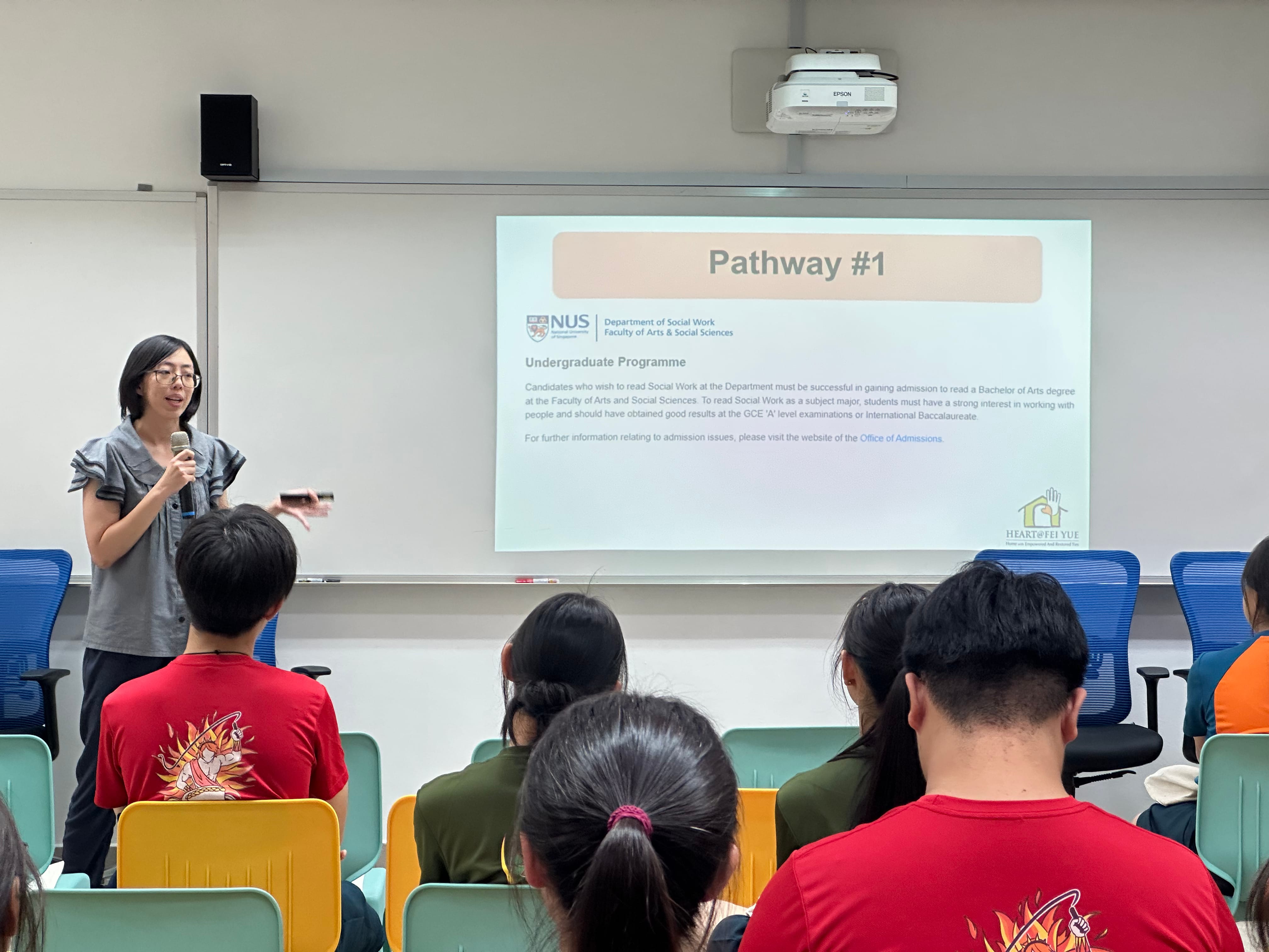 A woman speaks with a microphone before seated students, with a projected slide reading "Pathway #1" about NUS Social Work.