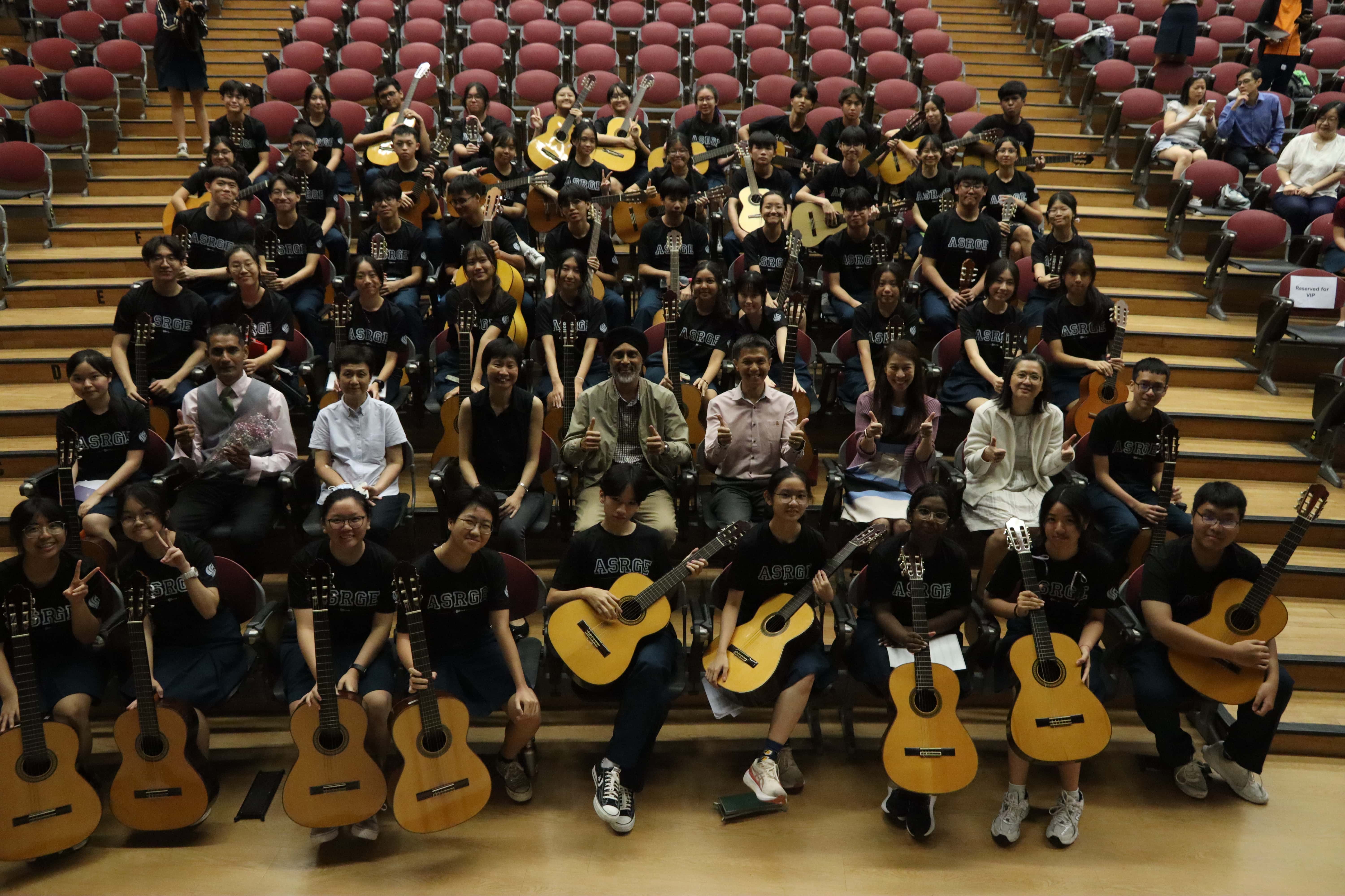Large group of students in black ASRGE shirts holding acoustic guitars, with the principal, vice-principals and teachers, posing together in a tiered auditorium.