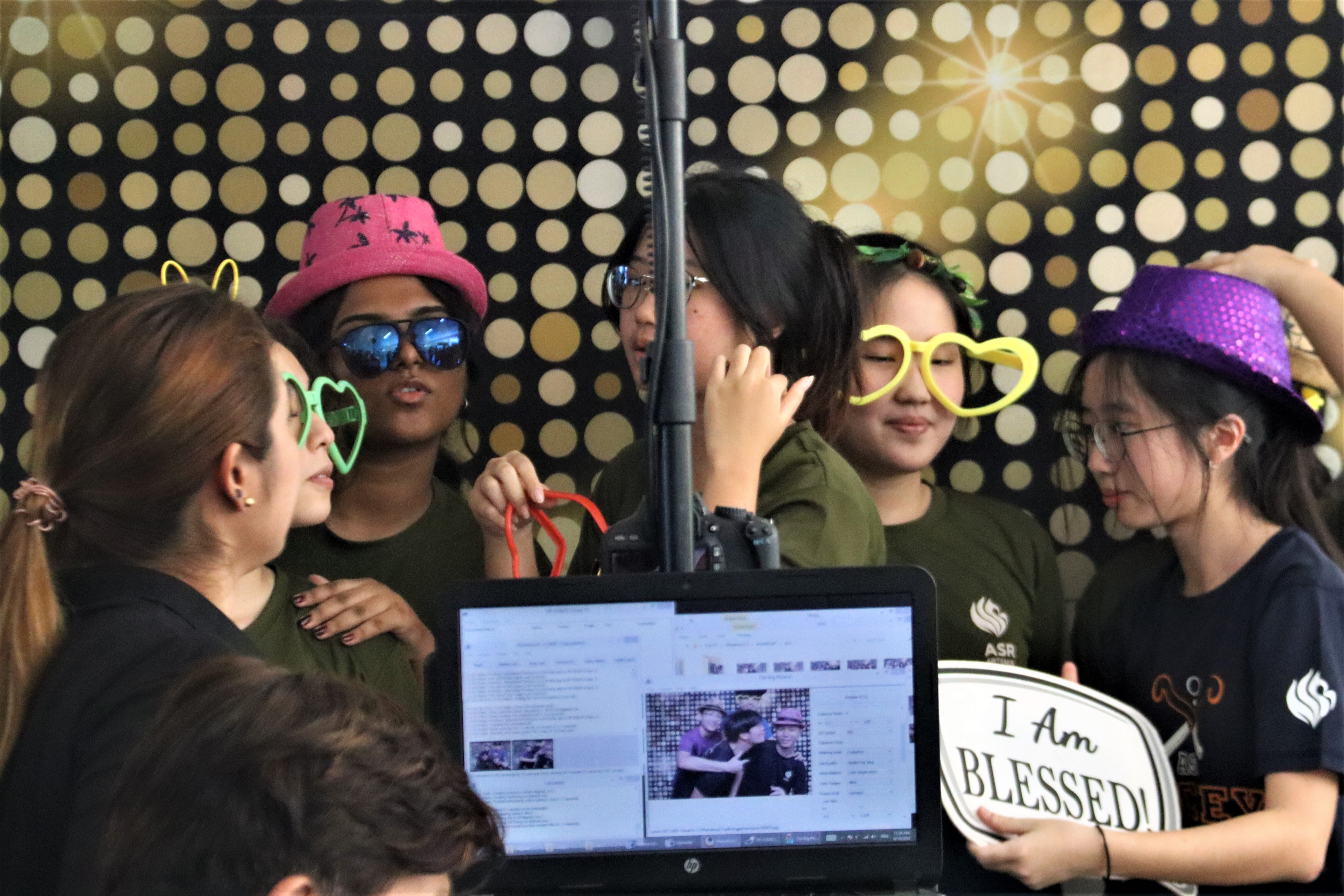 Group of young people at a photo booth wearing novelty glasses and hats, with a monitor and "I Am Blessed" sign visible.