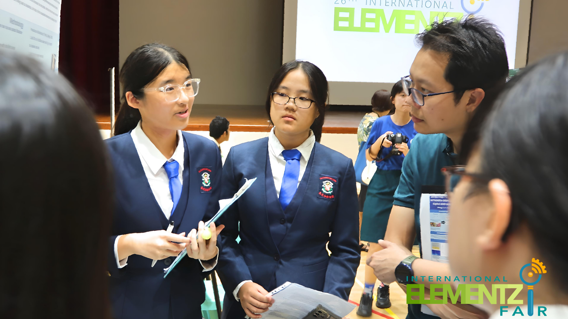 Two students in navy uniforms with blue ties speak with a judge at the International Elementz Fair.