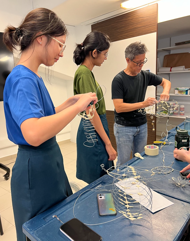 Three people wearing safety glasses work with wire sculptures on a blue worktable in an indoor workshop.