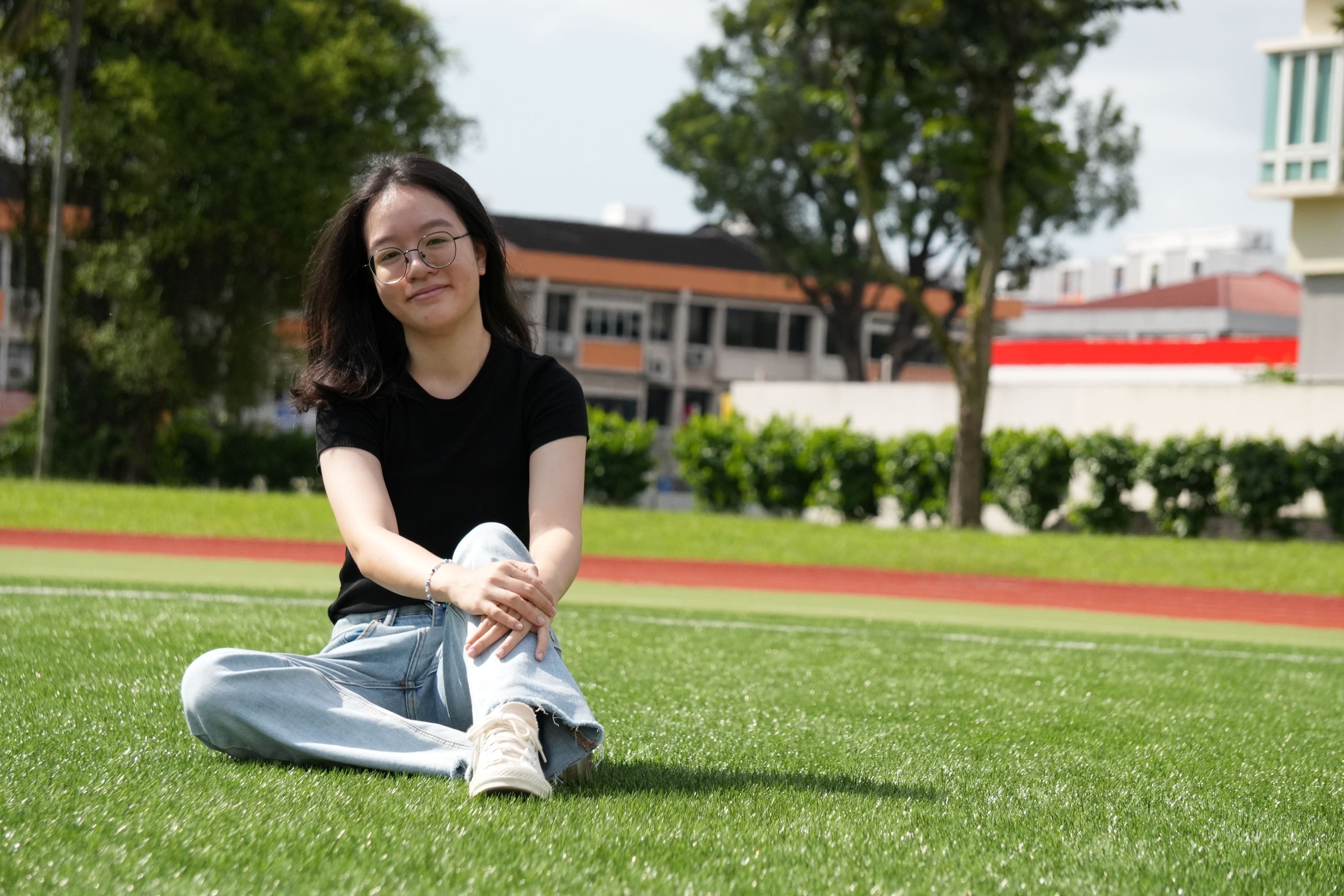Young woman with glasses in black shirt and jeans sits cross-legged on green grass near a running track.