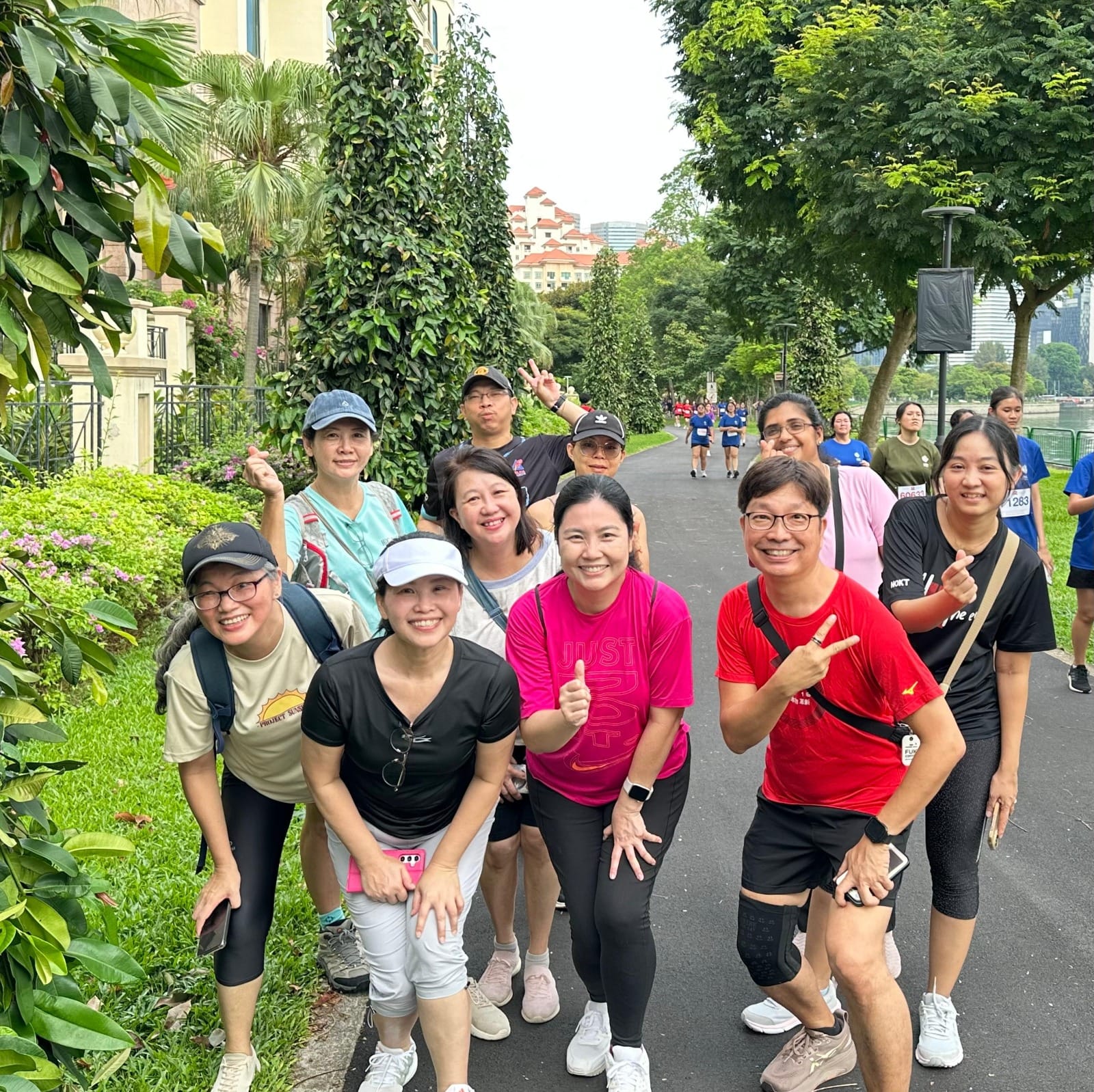 A group of smiling adults in athletic wear pose on a tree-lined path during an outdoor run or walk event.