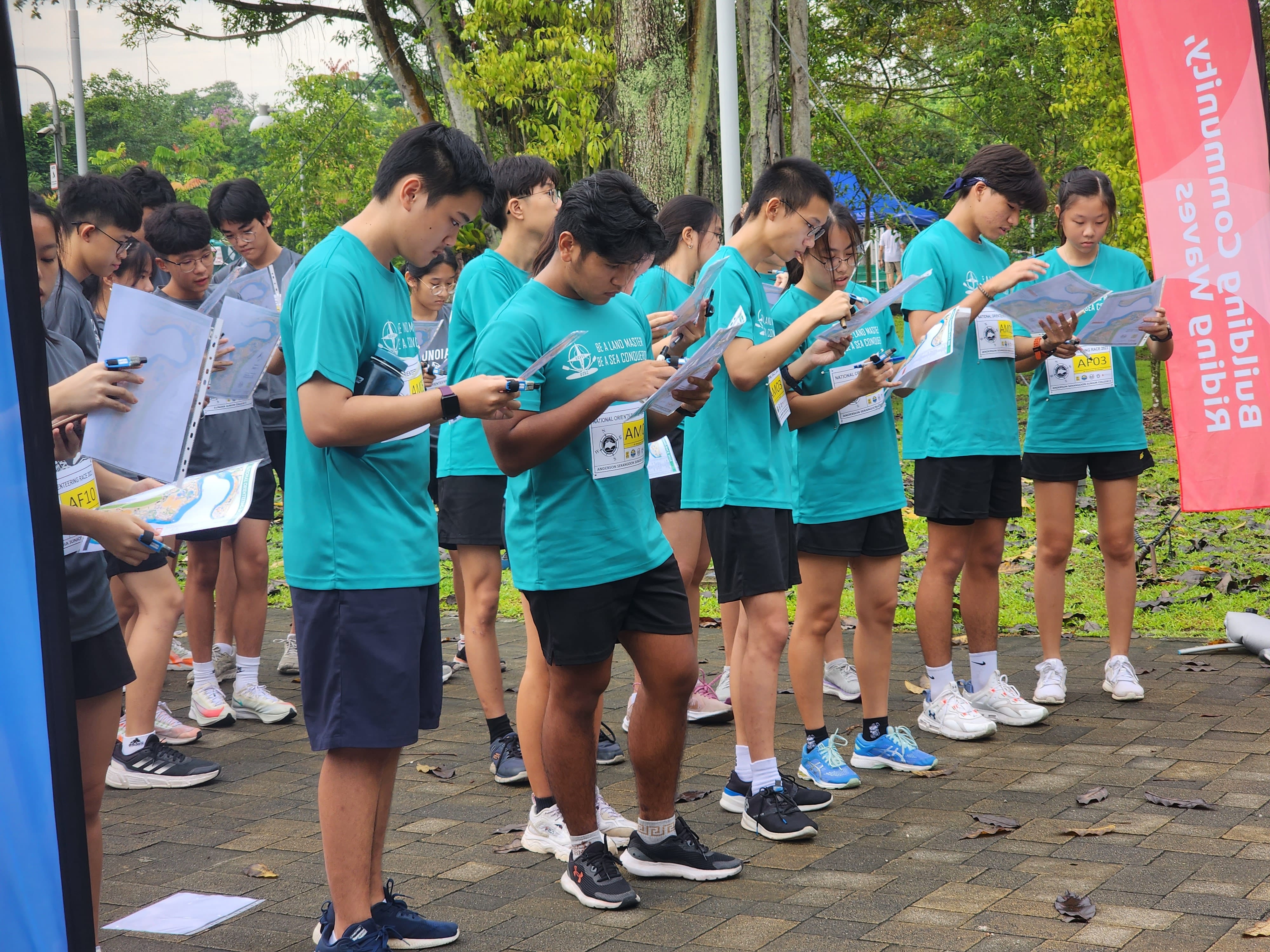A group of young people in teal shirts study maps outdoors near event banners and trees.