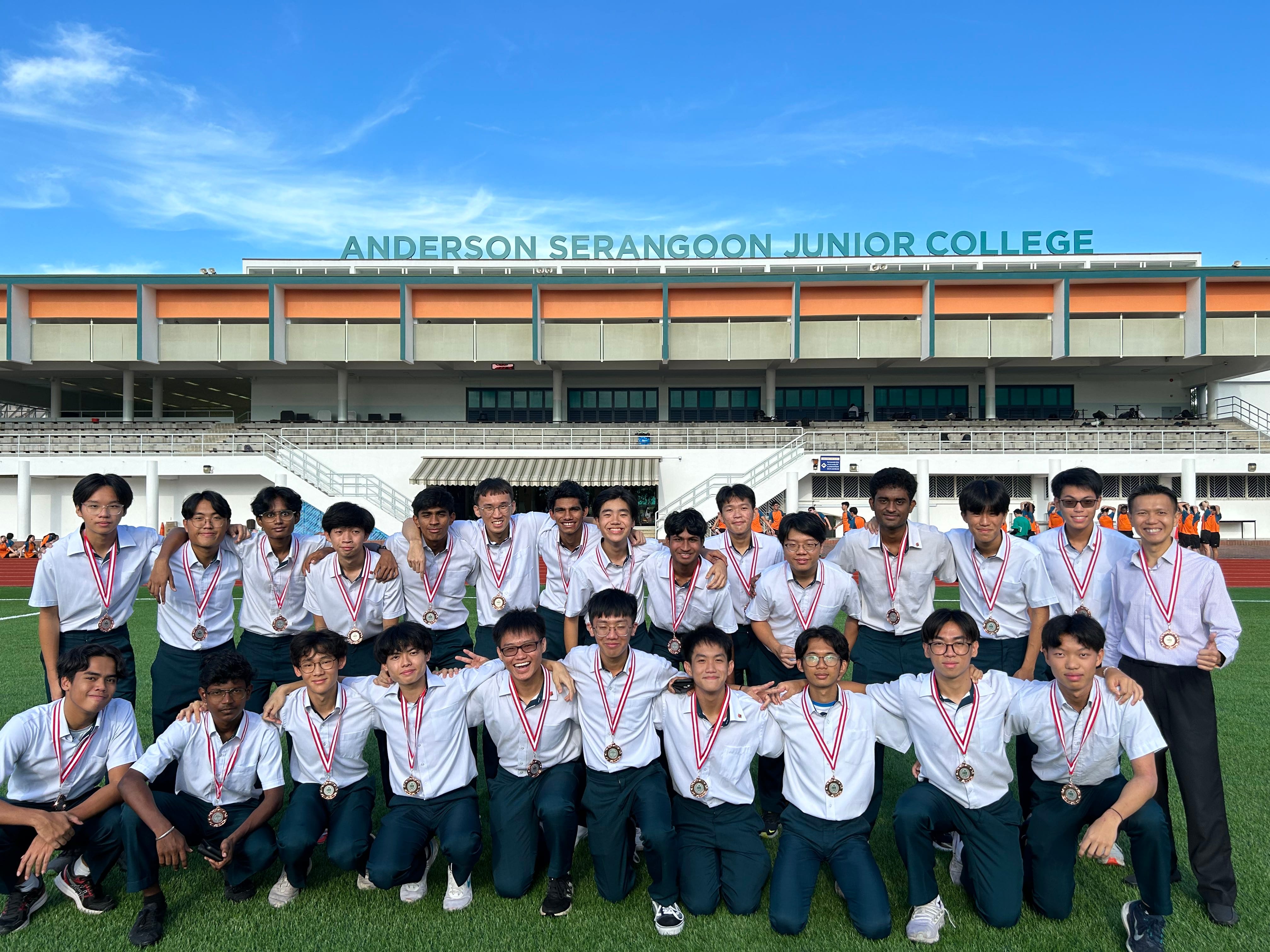 Group of young men in white shirts wearing medals posing on a field in front of Anderson Serangoon Junior College.