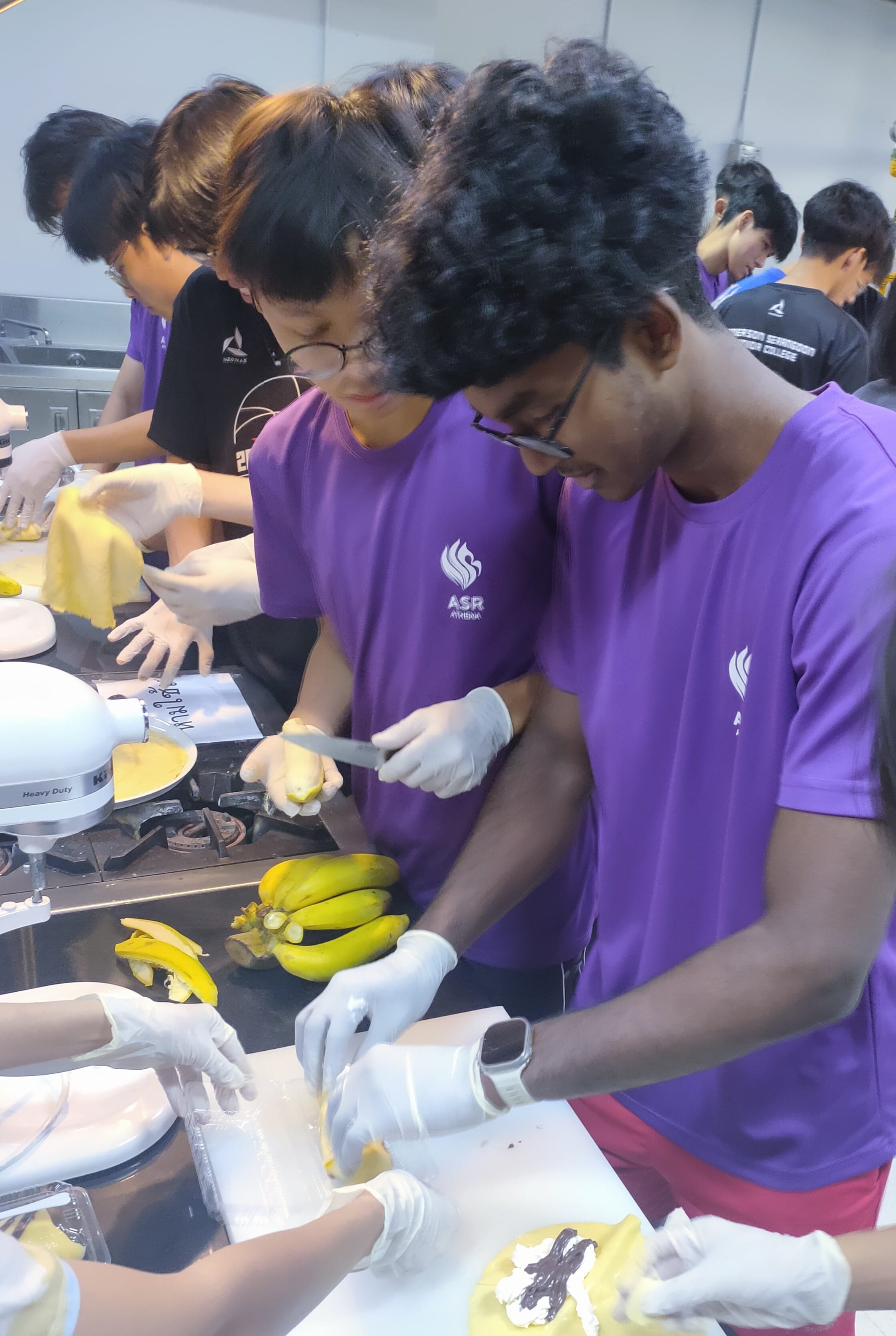 Two students in purple ASR shirts and gloves slice bananas on a counter during a group cooking activity.