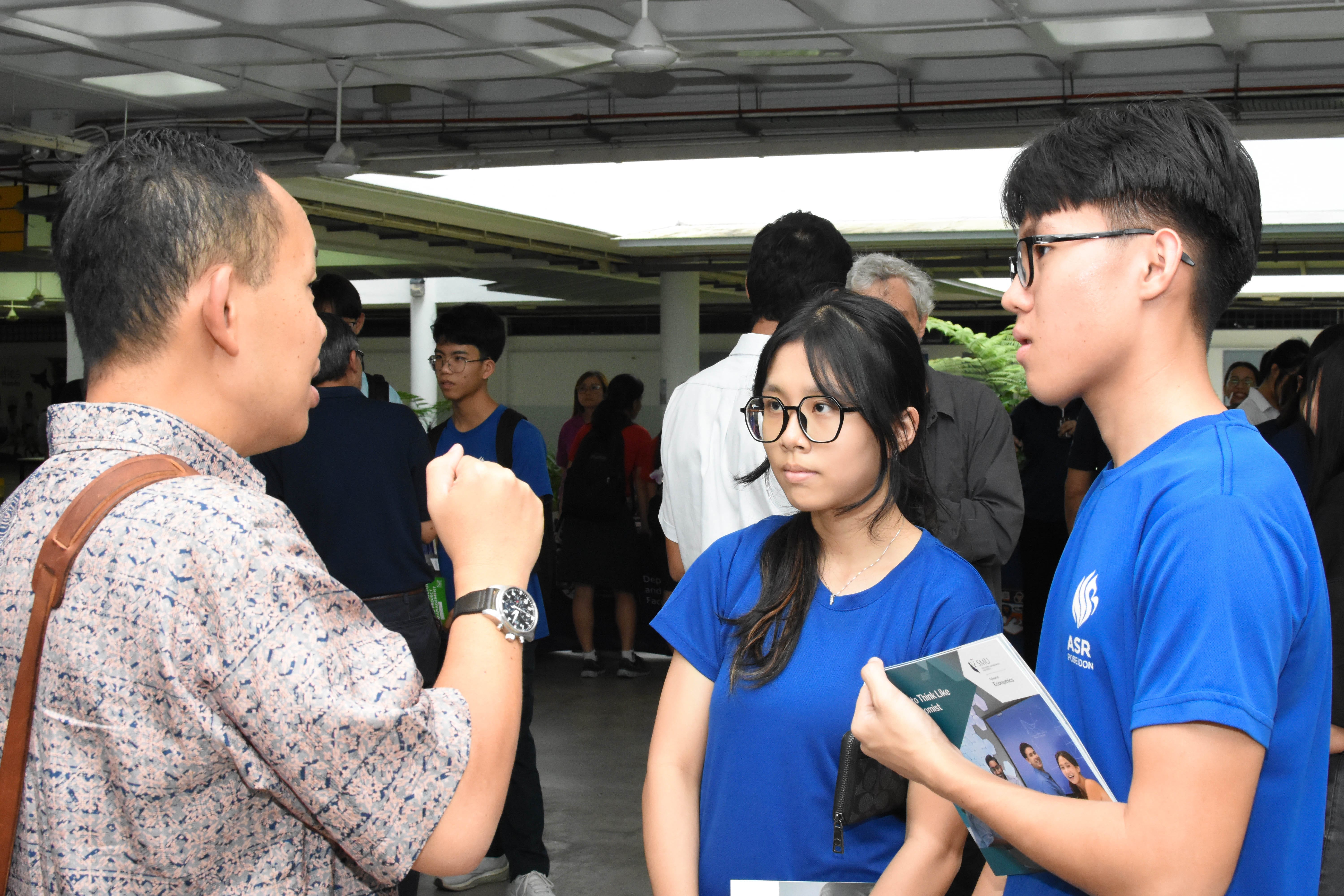A man talks to two students in blue shirts holding a brochure at a crowded indoor event.