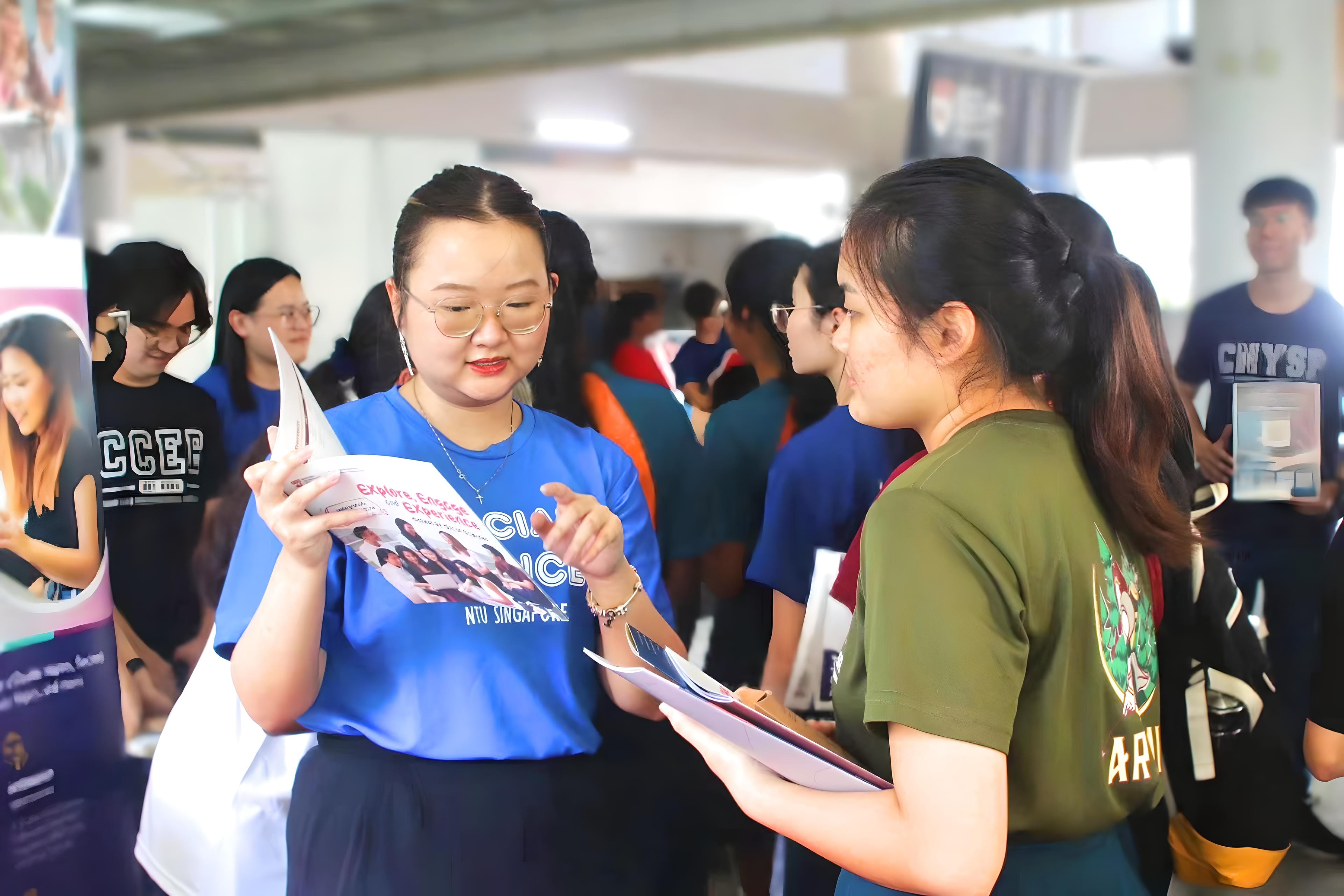 Two young women review a brochure together at a busy indoor fair, one wearing a blue NTU Singapore shirt.