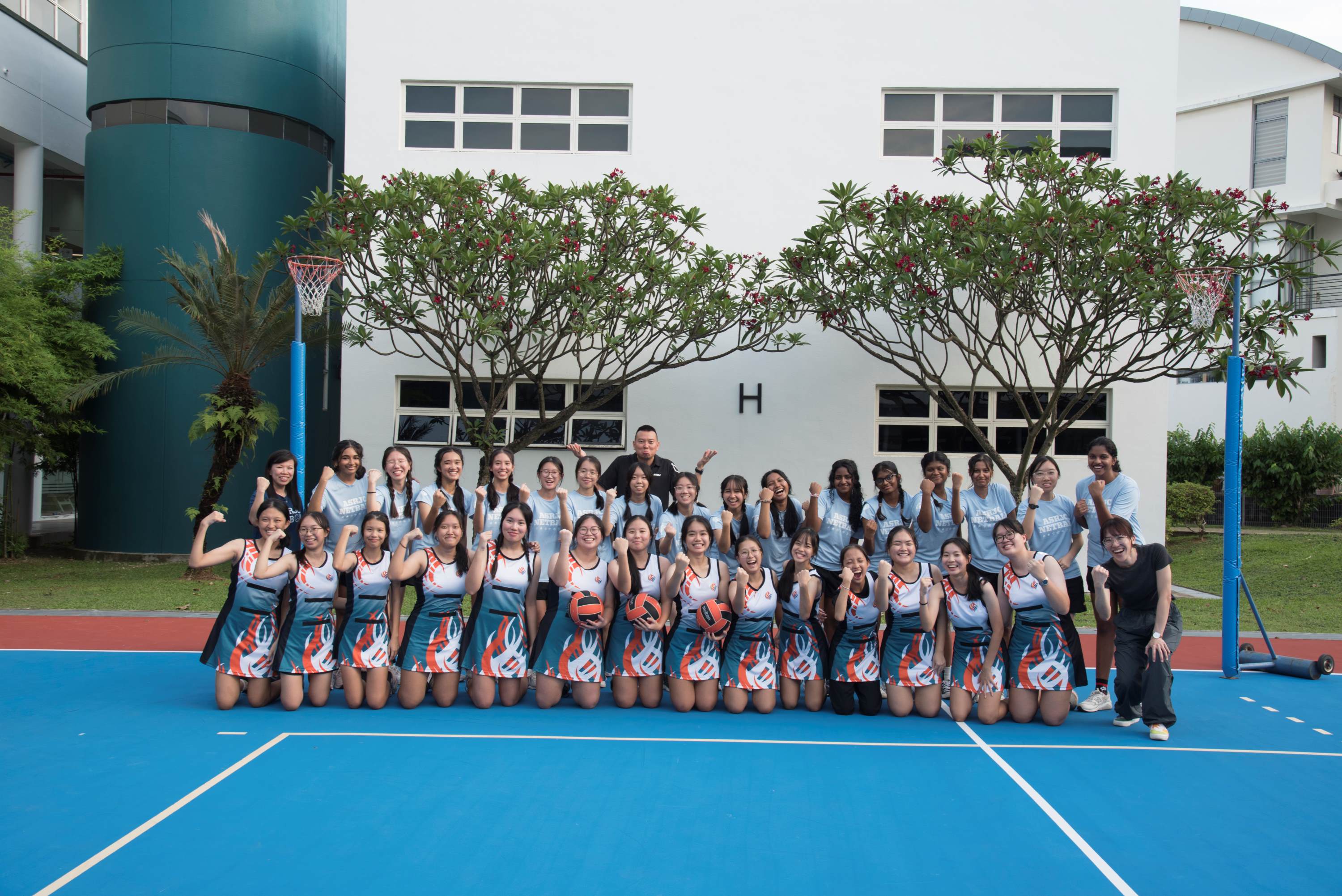A girls netball team poses on a blue court, wearing teal uniforms, holding balls, with netball posts and a school building behind.
