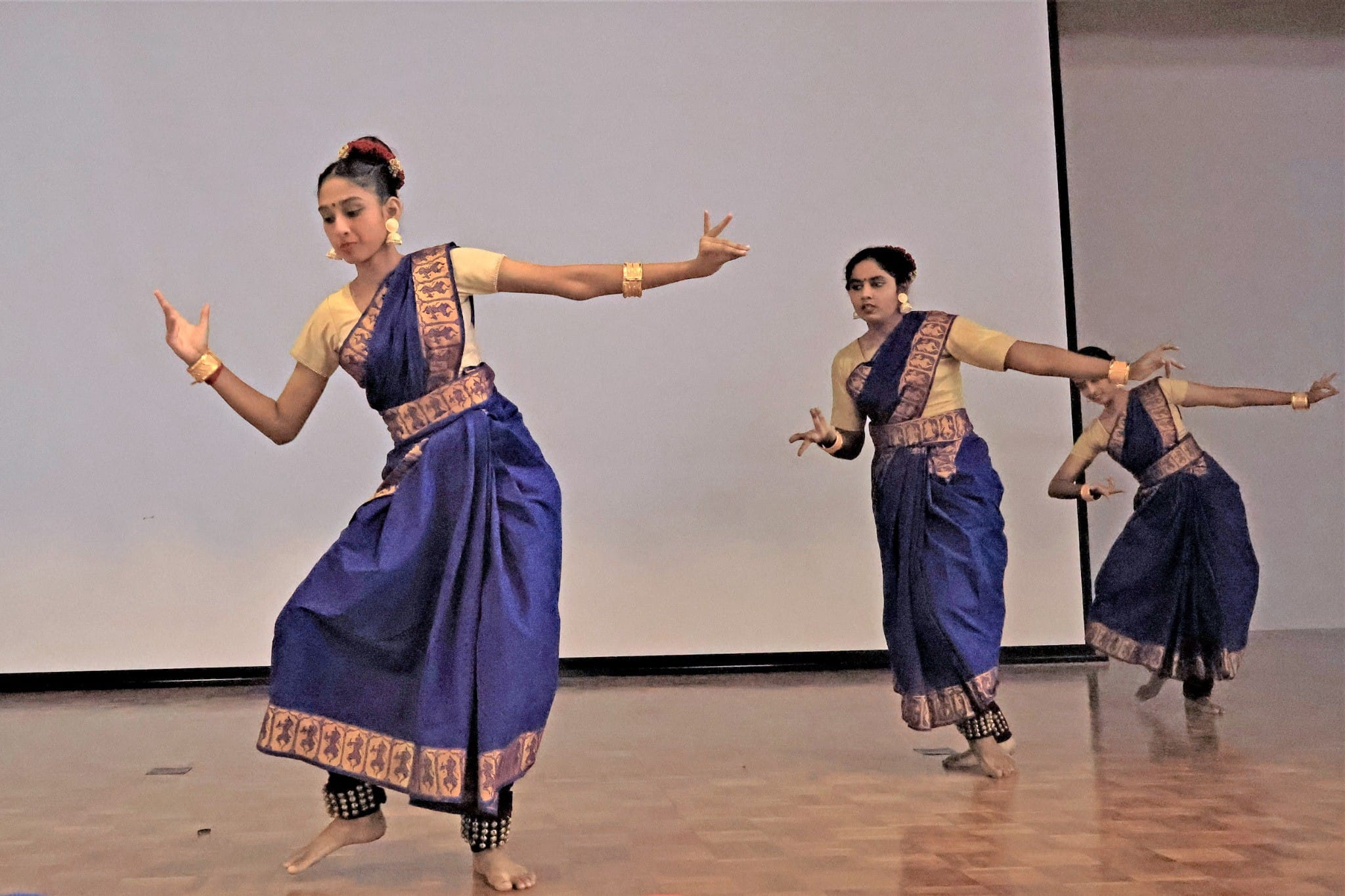 Three women in blue and gold sarees perform classical Indian dance on a wooden stage with arms gracefully extended.